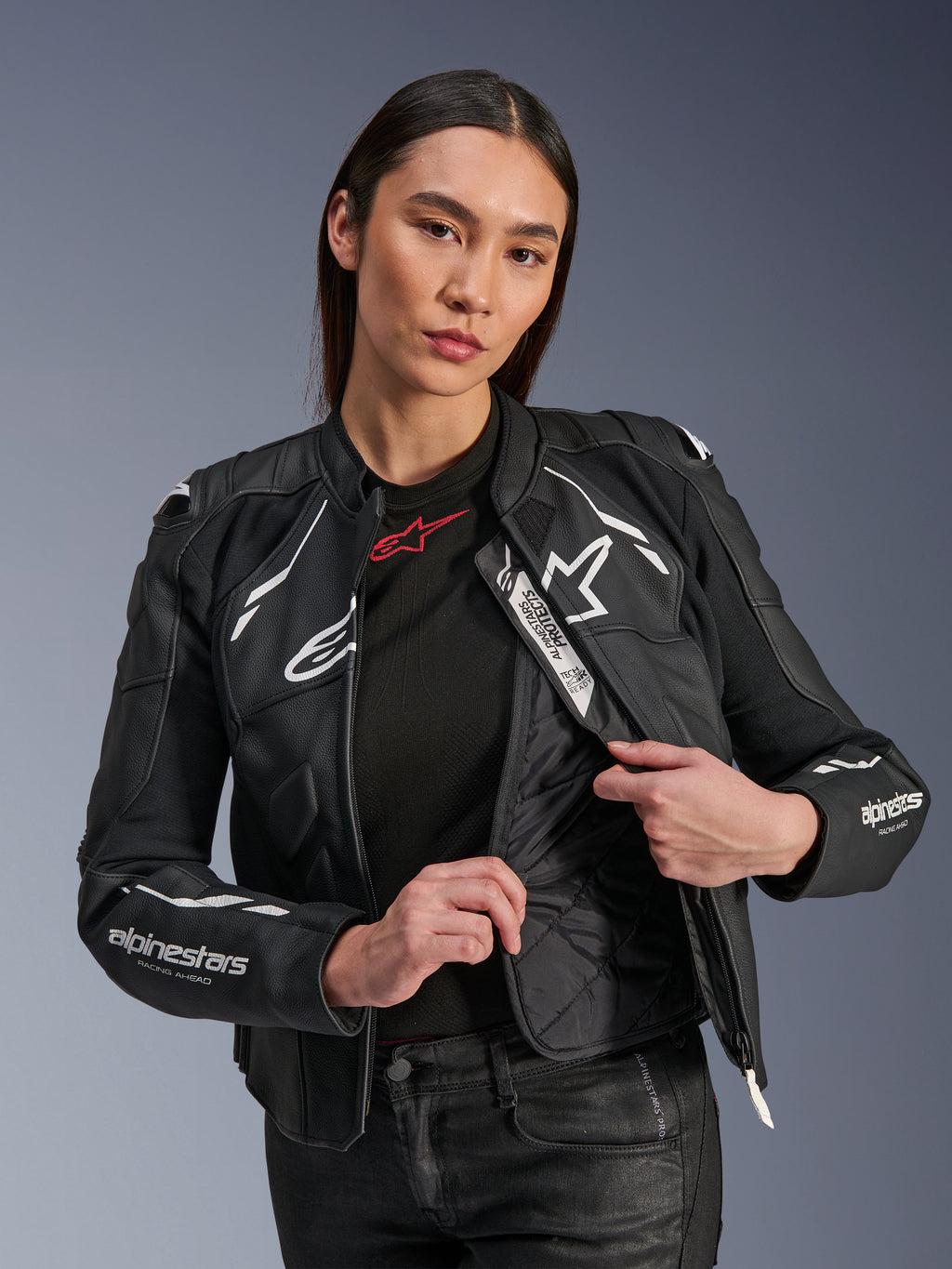 Modelka w damskiej kurtce skórzanej Alpinestars Stella Dusk w kolorze czarno-białym, prezentująca wewnętrzną pikowaną podszewkę termiczną i etykietę Tech-Air Ready, wykonana z wysokiej jakości skóry bydlęcej z białym logo gwiazdy i napisem „RACING AHEAD” na rękawach