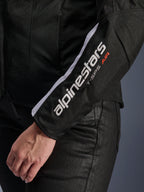 Alpinestars Stella T-SPS Air V2 Jacket, damska kurtka motocyklowa, czarno-biała, zbliżenie na rękaw z białym logo Alpinestars i napisem T-SPS AIR, siatkowa i tekstylna konstrukcja zapewniająca przepływ powietrza, noszona z czarnymi spodniami motocyklowymi