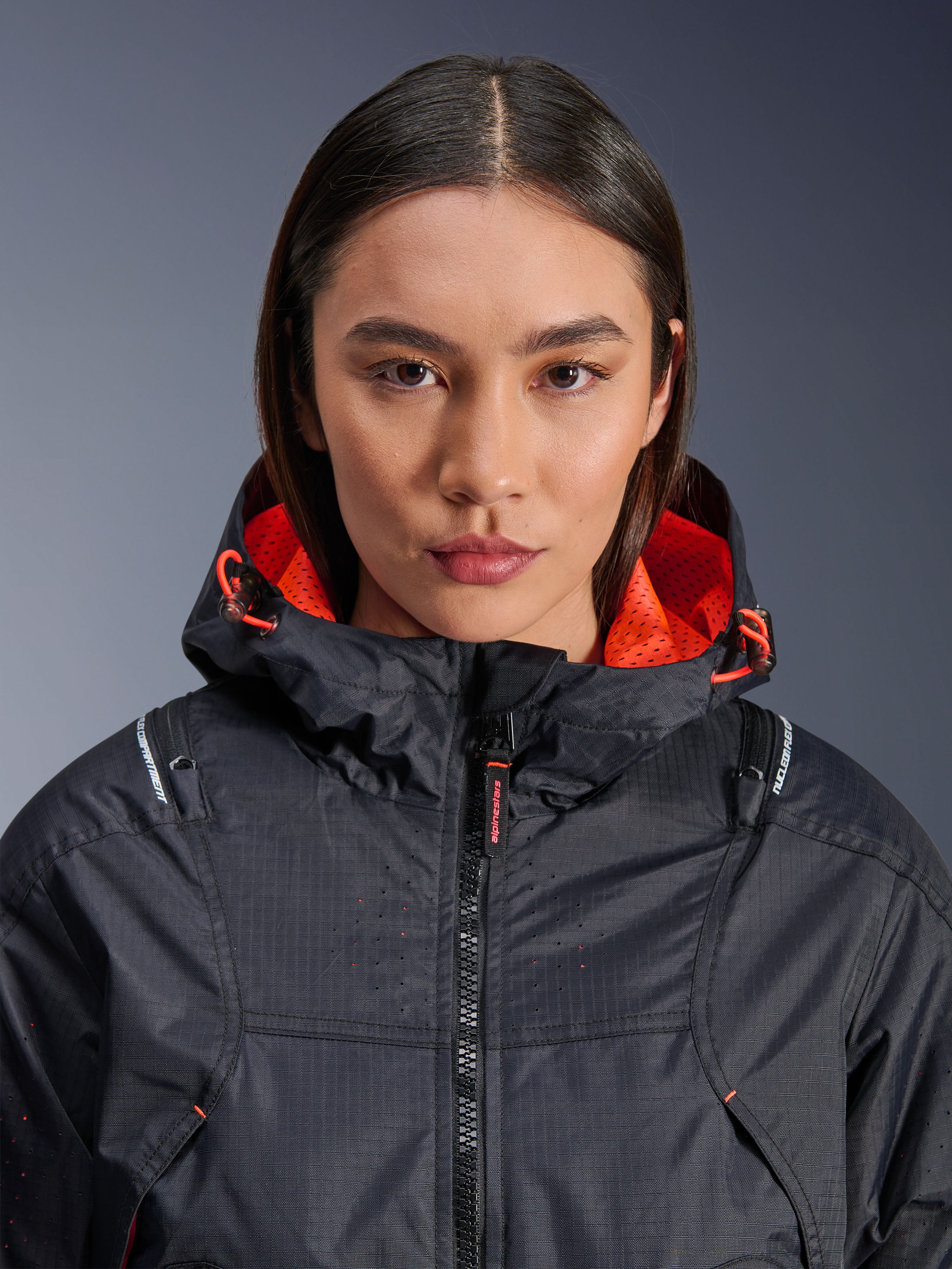 Kurtka Alpinestars Molly Air, damska kurtka motocyklowa, czarna z elementami w kolorze Fluo Coral, zbliżenie z przodu na modelce, materiał ripstop z laserową perforacją, jaskrawopomarańczowa siatkowa podszewka kaptura i ściągacze, z oznaczeniem ochraniaczy Nucleon Flex Plus