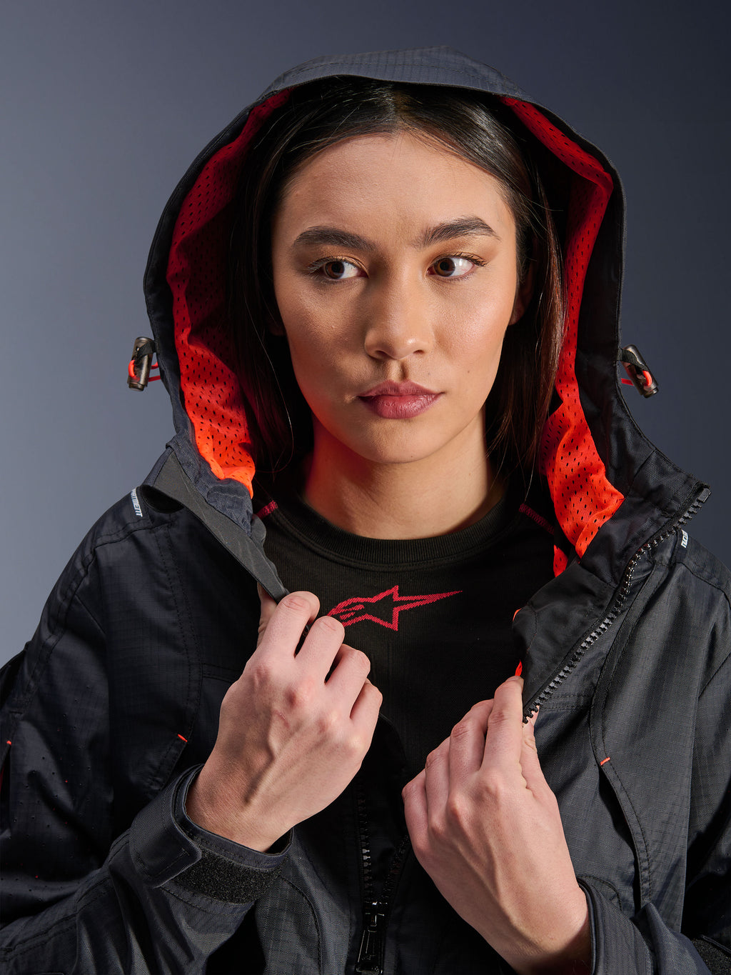 Alpinestars Molly Air Jacket, damska kurtka motocyklowa, czarna i koralowa fluo, modelka w założonym kapturze prezentująca jaskrawą koralową podszewkę z siateczki, tkanina tekstylna ripstop, nowoczesna miejska odzież motocyklowa