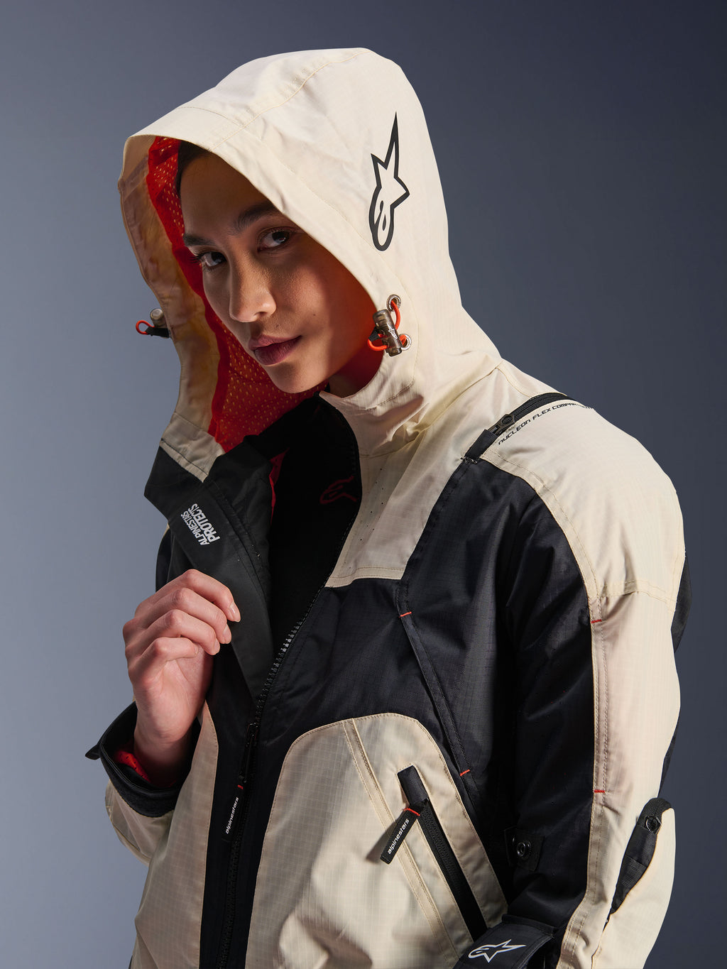 Alpinestars Molly Air Jacket w kolorze Black Almond, noszona przez modelkę z założonym kapturem, z kremowo-czarnymi panelami kolorystycznymi, wyrazistą pomarańczową podszewką z siateczki i technicznymi miejskimi elementami designu, w tym logo z gwiazdą i detalami z zamkami.