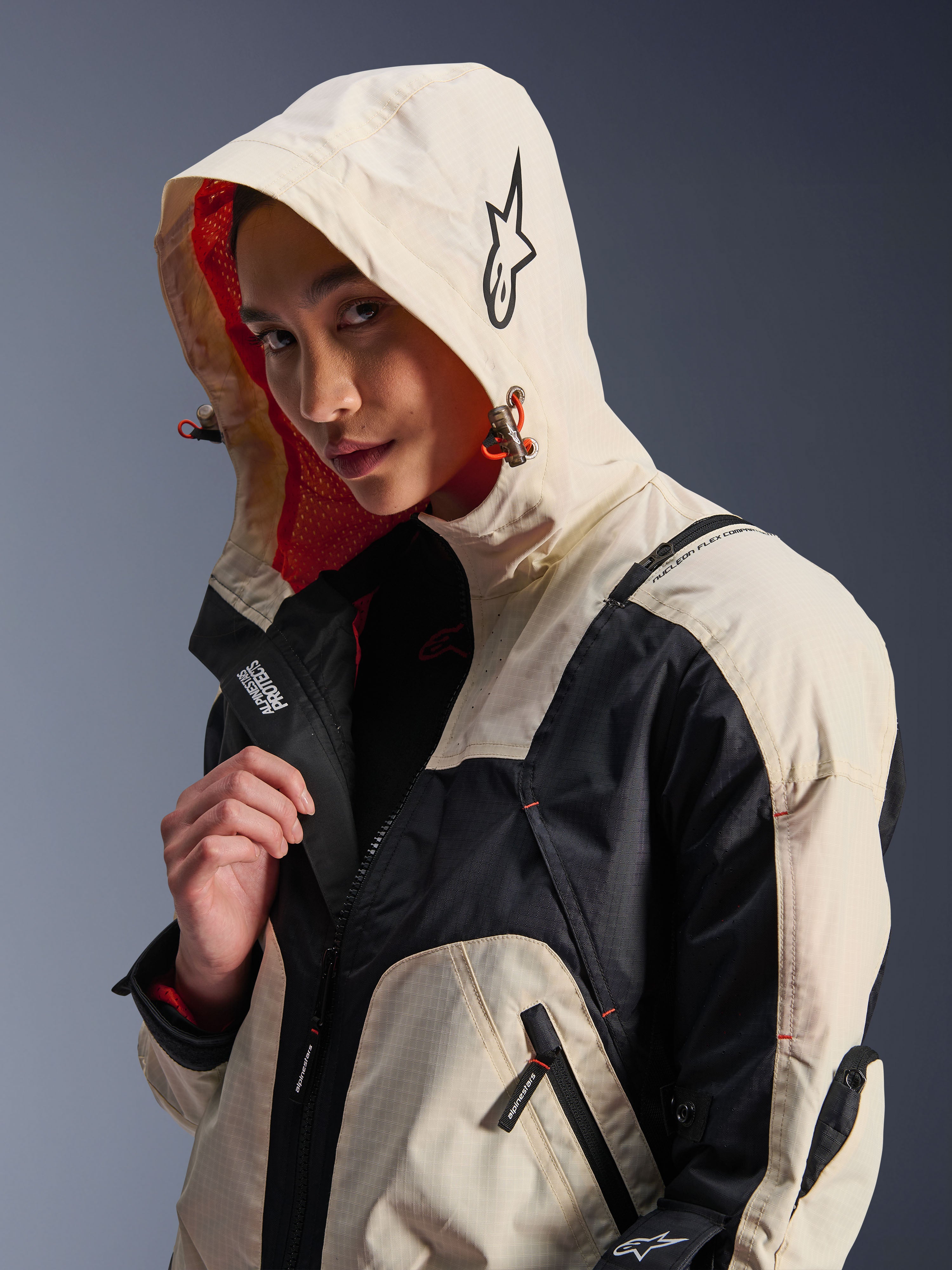 Alpinestars Molly Air Jacket w kolorze Black Almond, noszona przez modelkę z założonym kapturem, z kremowo-czarnymi panelami kolorystycznymi, wyrazistą pomarańczową podszewką z siateczki i technicznymi miejskimi elementami designu, w tym logo z gwiazdą i detalami z zamkami.
