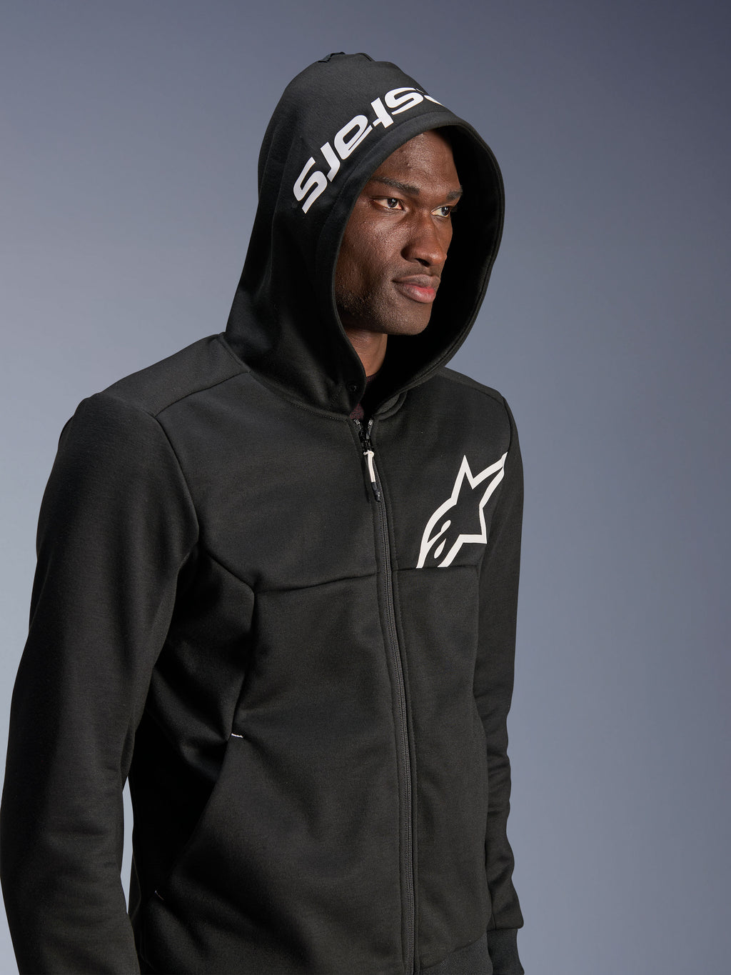 Alpinestars Chrome V2 Sport Hoodie, czarna miejska bluza motocyklowa noszona przez modela z założonym kapturem, białe logo gwiazdy na klatce piersiowej i biały napis stars na kapturze, konstrukcja z technicznego polaru, casualowy motocyklowy streetwear