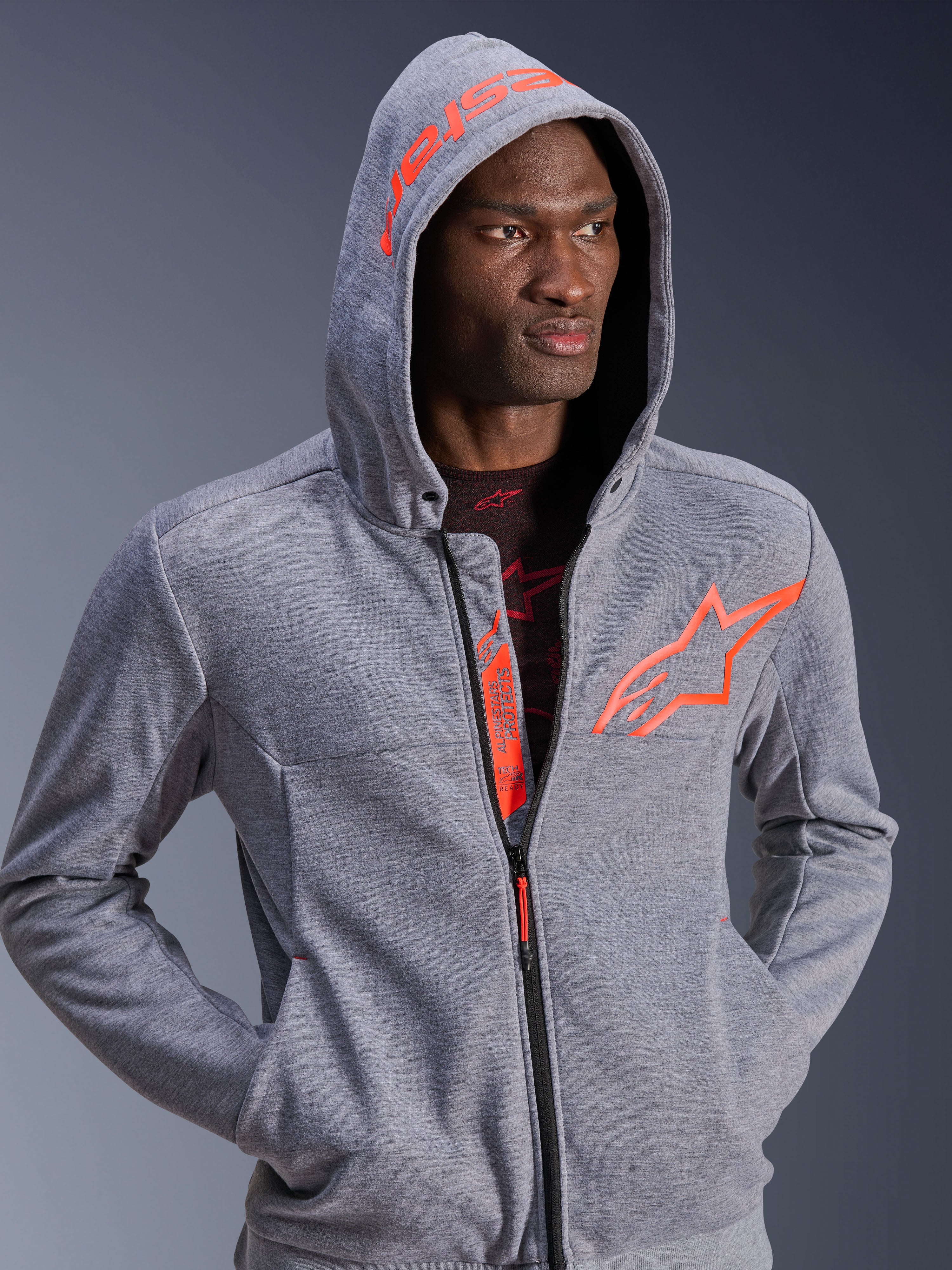 Alpinestars Chrome V2 Sport Hoodie, motocyklowa bluza z kapturem, szary melanż i czerwony fluo, mężczyzna z założonym kapturem, duże pomarańczowe logo gwiazdy na piersi i napis Alpinestars na kapturze, casualowy miejski styl