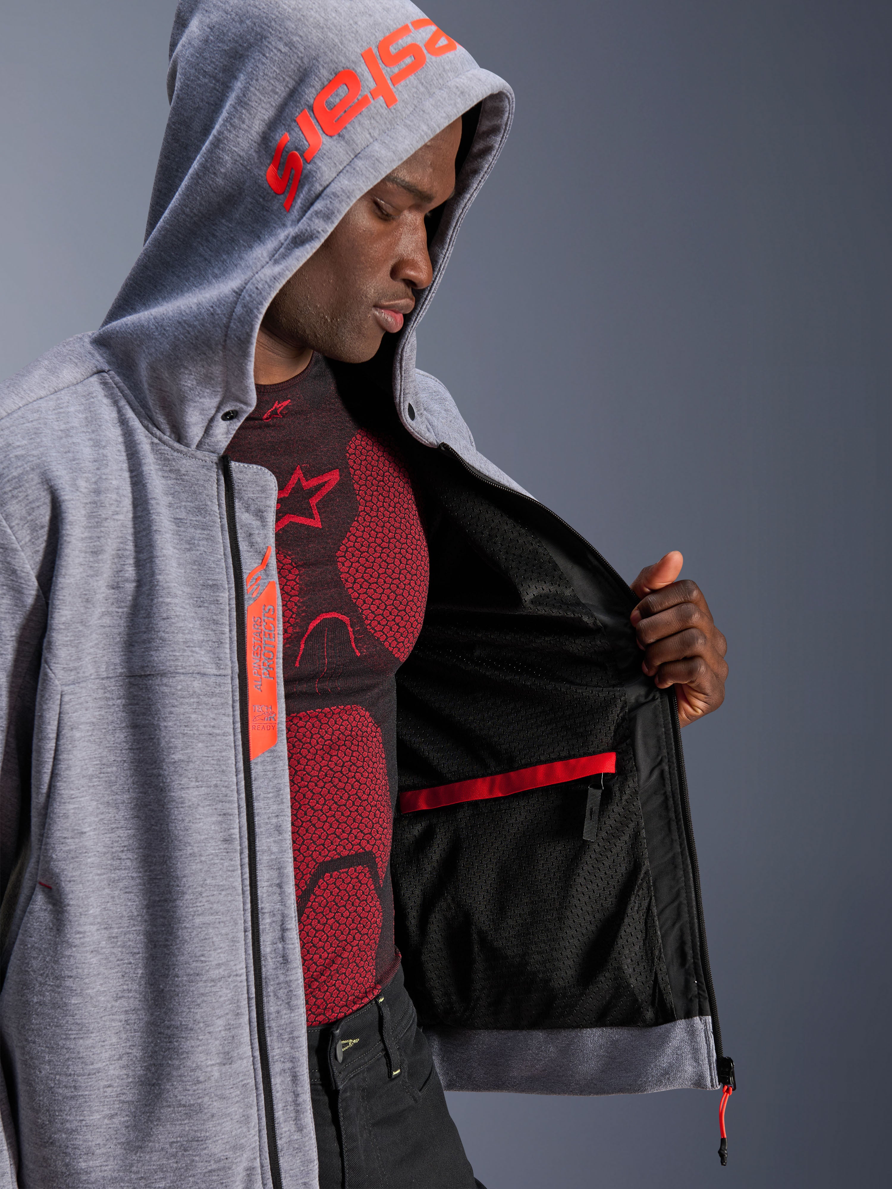 Alpinestars Chrome V2 Sport Hoodie, bluza motocyklowa, szary melanż i czerwony fluo, męski model prezentujący wewnętrzną czarną podszewkę z siatki i czerwoną wodoodporną kieszeń, czerwone logo na kapturze, etykieta Tech-Air Ready, sportowa sylwetka do jazdy miejskiej