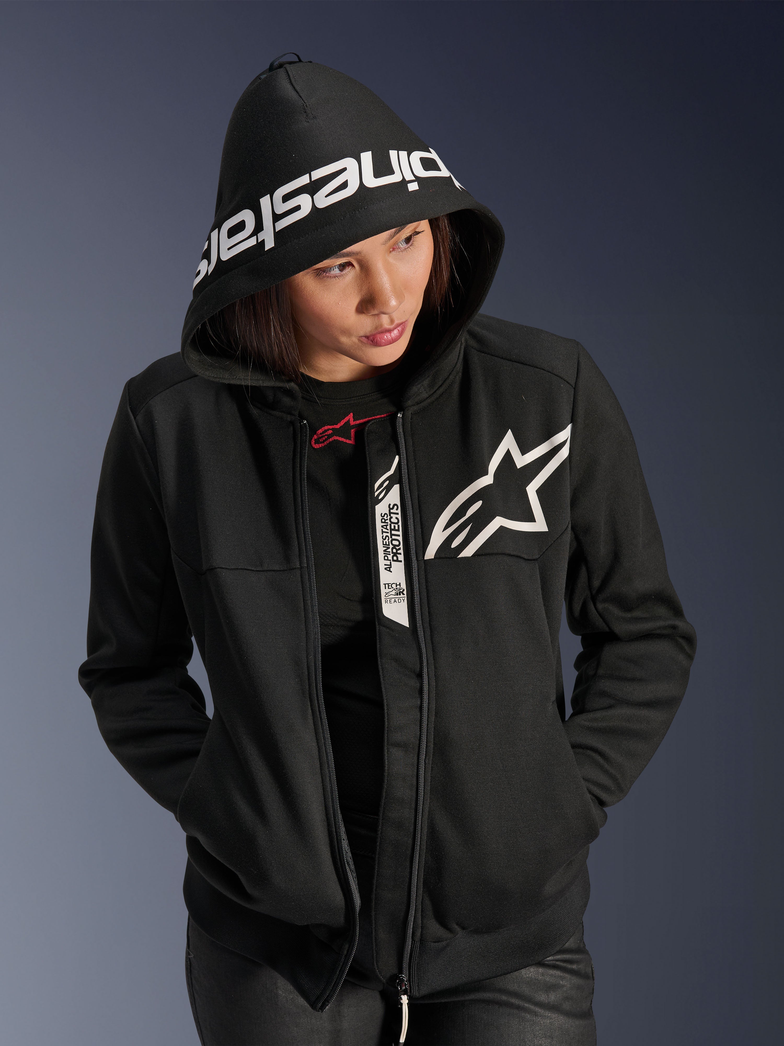 Alpinestars Stella Chrome V2 Sport Hoodie, damska bluza motocyklowa, czarno-biała, białe logo gwiazdy na piersi, branding Alpinestars na krawędzi kaptura, kobieta z założonym kapturem, swobodny miejski styl sportowy, detale Tech-Air Ready