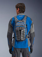 Alpinestars AT-15 Back Pack, przygodowy plecak motocyklowy, ciemnoszary i grafitowy, noszony przez motocyklistę na niebiesko-szarej kurtce, z logo All Terra, ergonomiczna konstrukcja strukturalna, regulowane paski i niebieskie akcenty przy zamkach.