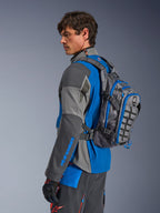 Alpinestars AT-15 Back Pack, przygodowy plecak motocyklowy, ciemnoszary z niebieskimi akcentami, noszony przez motocyklistę w dopasowanej szaro-niebieskiej technicznej odzieży motocyklowej, wyposażony w panel MOLLE i wytrzymałą konstrukcję do jazdy w terenie.