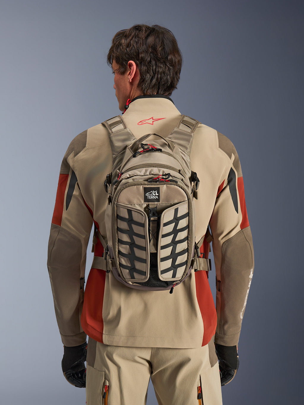 Alpinestars AT-15 Back Pack, Aluminum Walnut, techniczny plecak motocyklowy adventure noszony przez motocyklistę w beżowo-czerwonej kurtce, ze wzorem bieżnika opony, logo TERRA, czerwonymi uchwytami suwaków i regulowanymi paskami