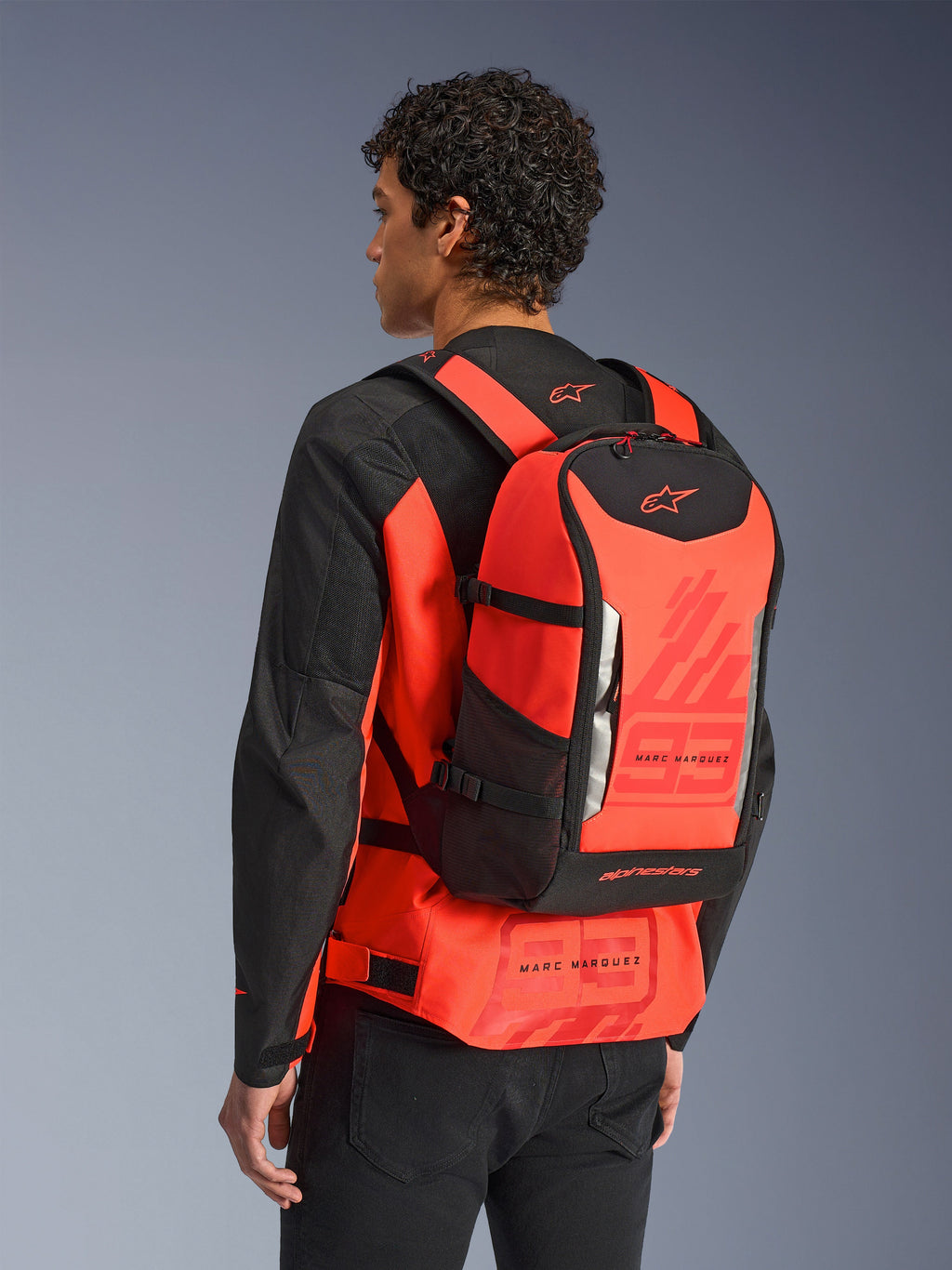 Plecak Alpinestars MM93 AMP3, techniczny plecak motocyklowy, czerwony fluo i czarny, model widoczny od tyłu w pasującej czerwono-czarnej kurtce, grafiki Marca Marqueza „93”, regulowane paski, nowoczesny sportowy design.