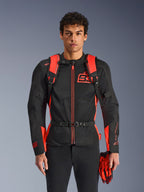 Mężczyzna w plecaku Alpinestars MM93 AMP3, kolor czerwony fluo i czarny, wyposażony w regulowane paski na ramiona i pasek piersiowy z klamrą, w stylizacji z dopasowaną kurtką motocyklową i rękawicami MM93, odzież techniczna z kolekcji Marca Marqueza