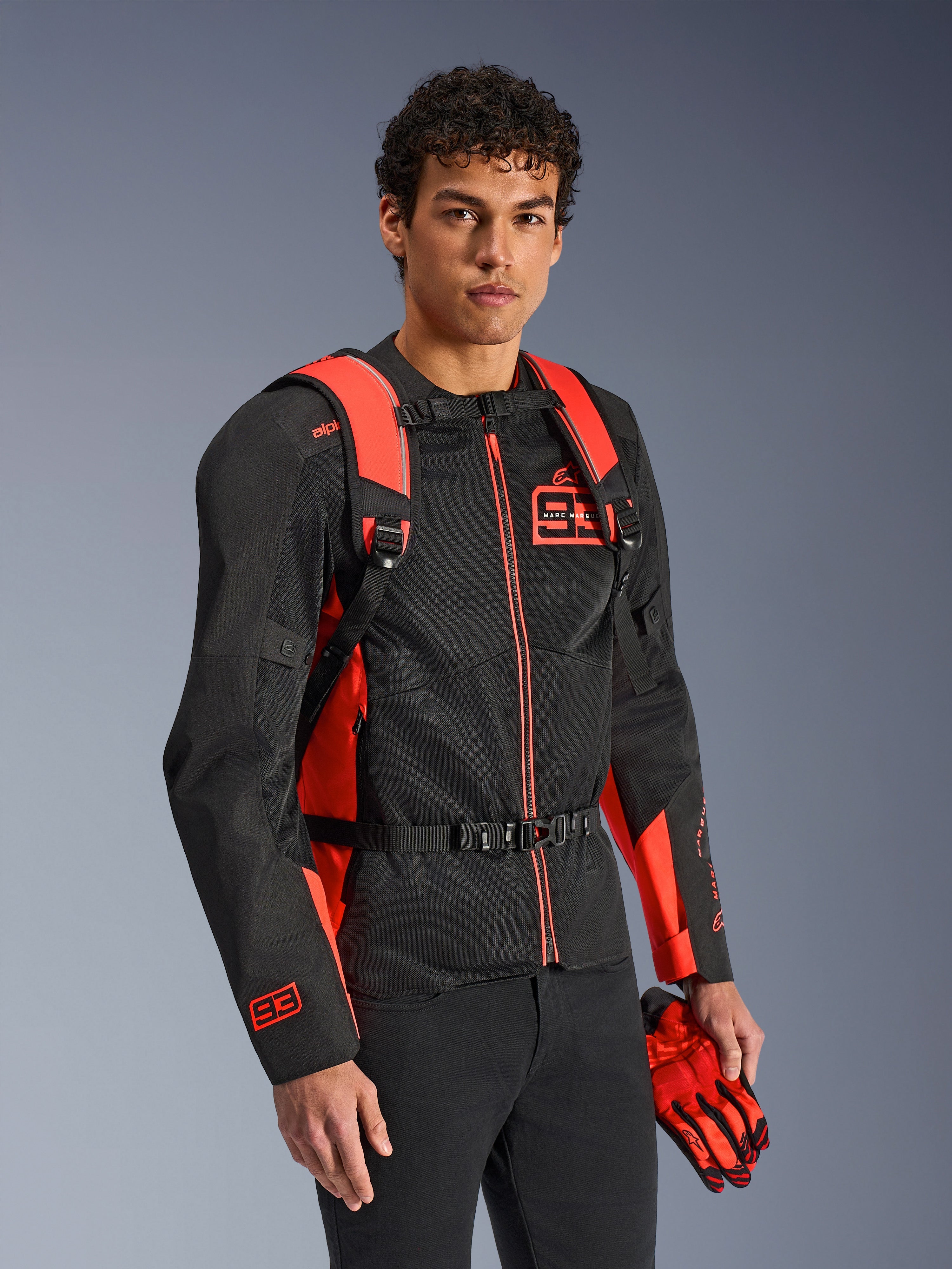 Plecak Alpinestars MM93 AMP3, jaskrawoczerwony i czarny, noszony przez modela z pasującą czarno-czerwoną kurtką motocyklową i rękawicami MM93, widok z przodu podkreślający regulowane paski naramienne i klamrę piersiową, techniczny sprzęt inspirowany wyścigami