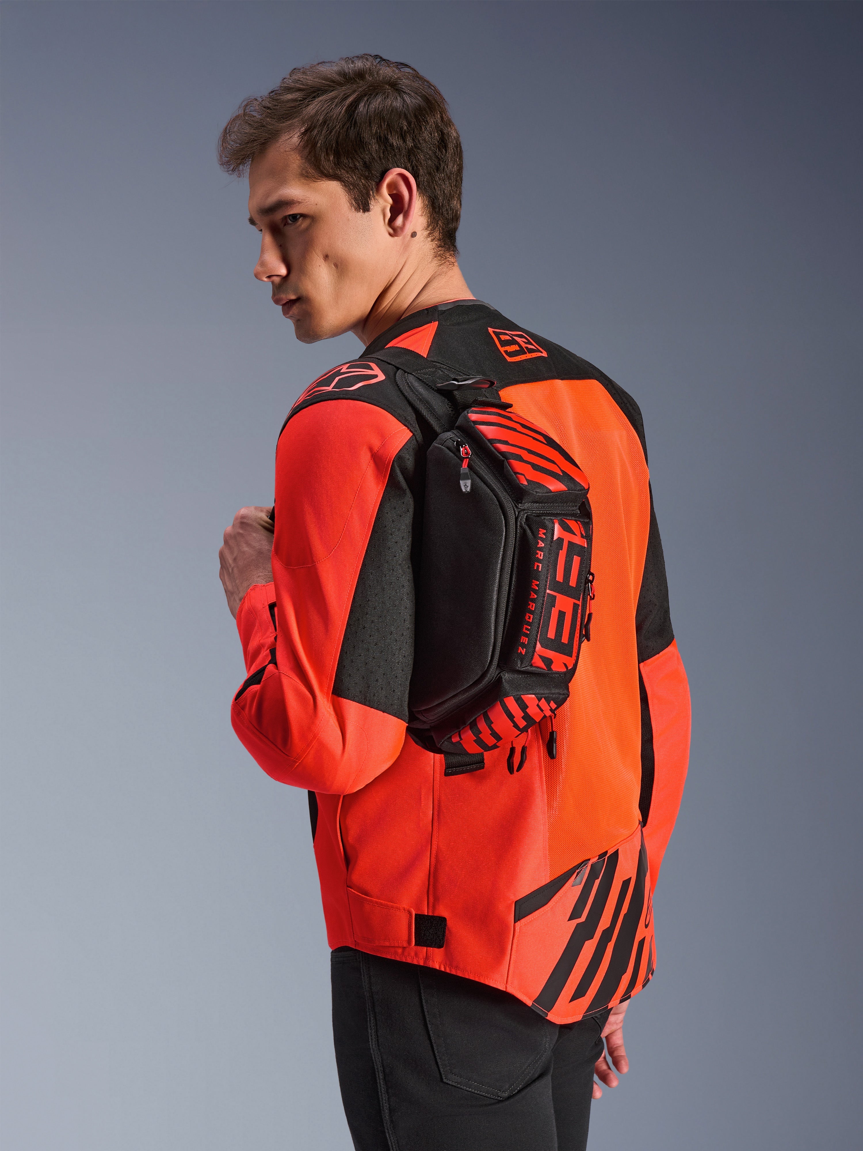 Torba na pas Alpinestars MM93, czarna z jaskrawoczerwonymi (Red Fluo) i jasnoczerwonymi (Bright Red) elementami, noszona przez modela na plecach na czerwono-czarnej kurtce motocyklowej, z wyraźnym brandingiem MM93 i grafikami Marca Marqueza