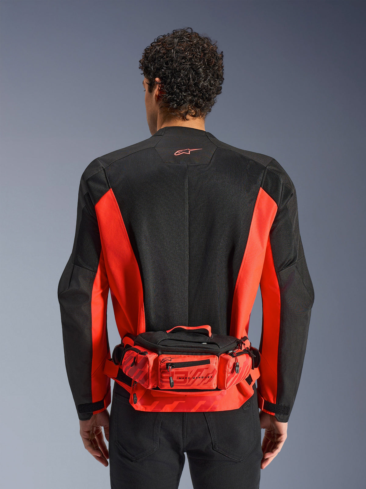 Alpinestars MM93 Waist Bag, czerwono-czarna torba na pas, noszona na czarno-czerwonej kurtce motocyklowej, z logo Marc Marquez 93, regulowany pasek i kieszenie zapinane na zamek, funkcjonalne akcesorium inspirowane wyścigami