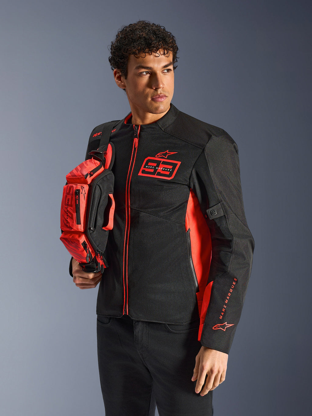 Alpinestars MM93 Waist Bag, torba na pas w kolorze czerwonym fluo i czarnym, trzymana przez modela w pasującej czarno-czerwonej kurtce motocyklowej, wyposażona w wiele przegród na zamek i branding Marca Marqueza, praktyczne wyposażenie do jazdy sportowej