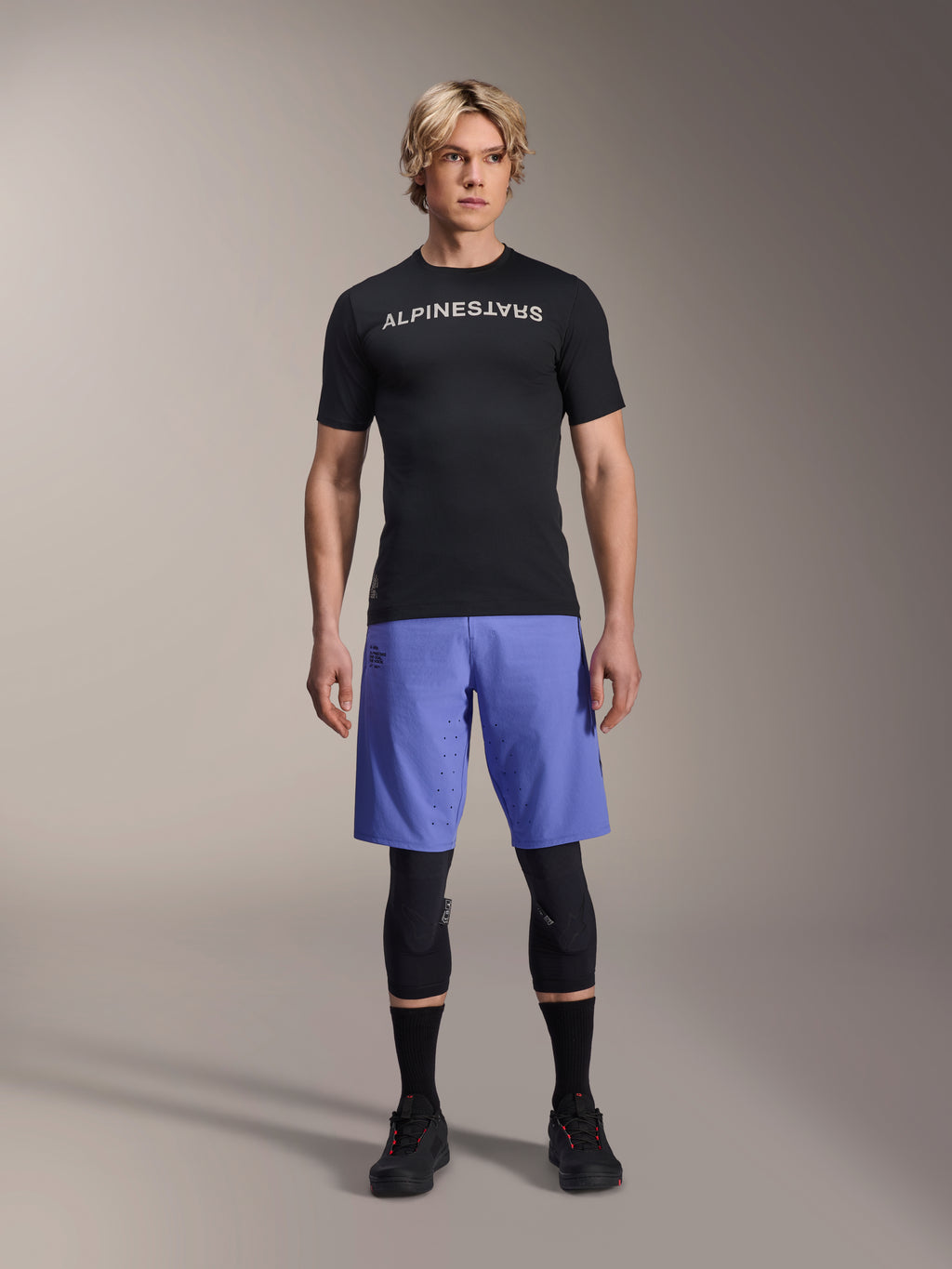 Koszulka Alpinestars A-Aria Switch, koszulka MTB z krótkim rękawem, czarna z białym lustrzanym logo Alpinestars, noszona przez modela w niebieskich spodenkach rowerowych i czarnych ochraniaczach na kolana, oddychająca odzież techniczna do jazdy po szlakach