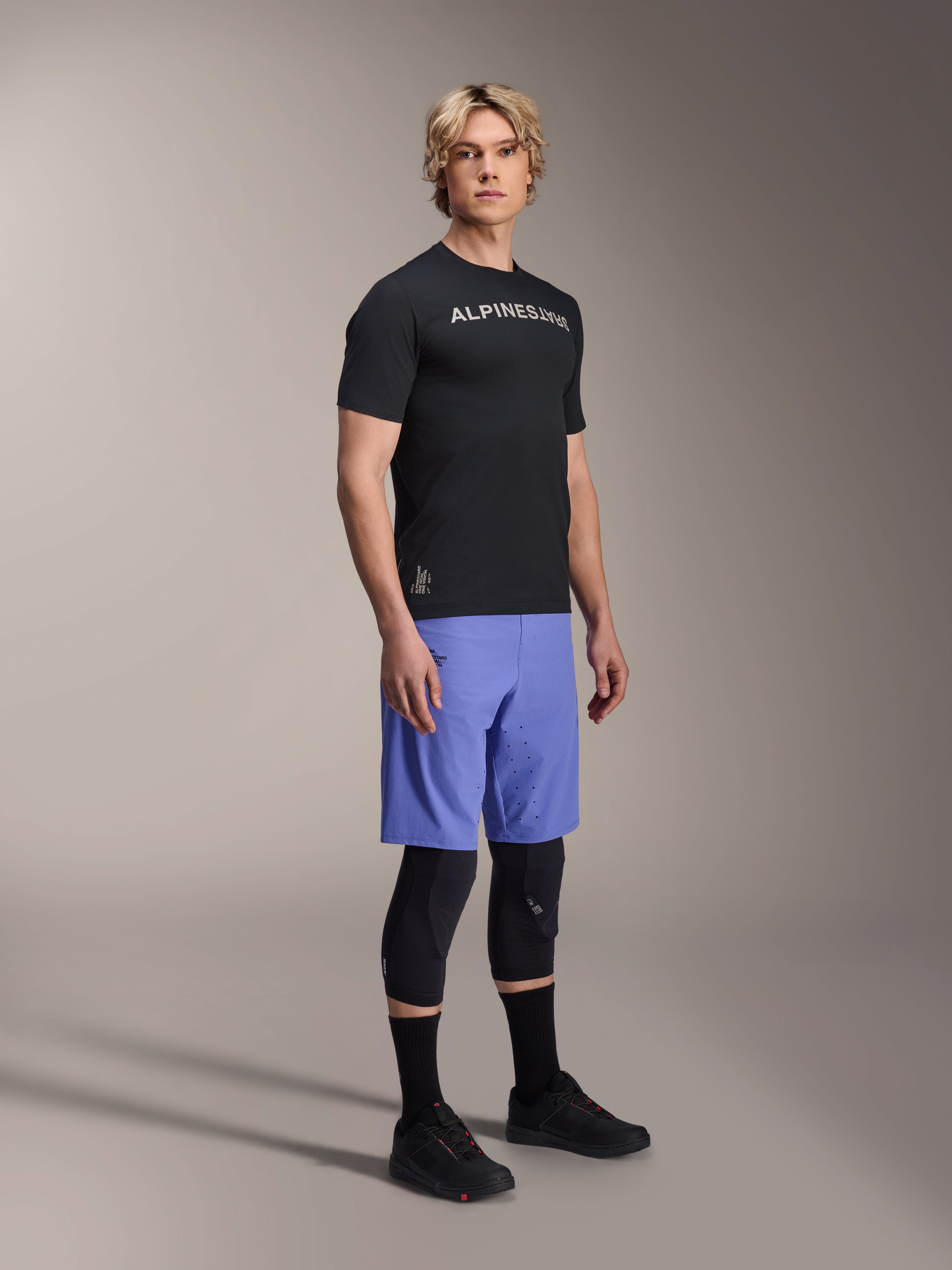 Male model wearing Alpinestars mountain bike apparel, featuring the black A-Aria Switch SS Jersey and Baja Blue A-Aria Lite Shorts, paired with black knee guards, black socks, and black mountain bike shoes in a full-body studio shot.