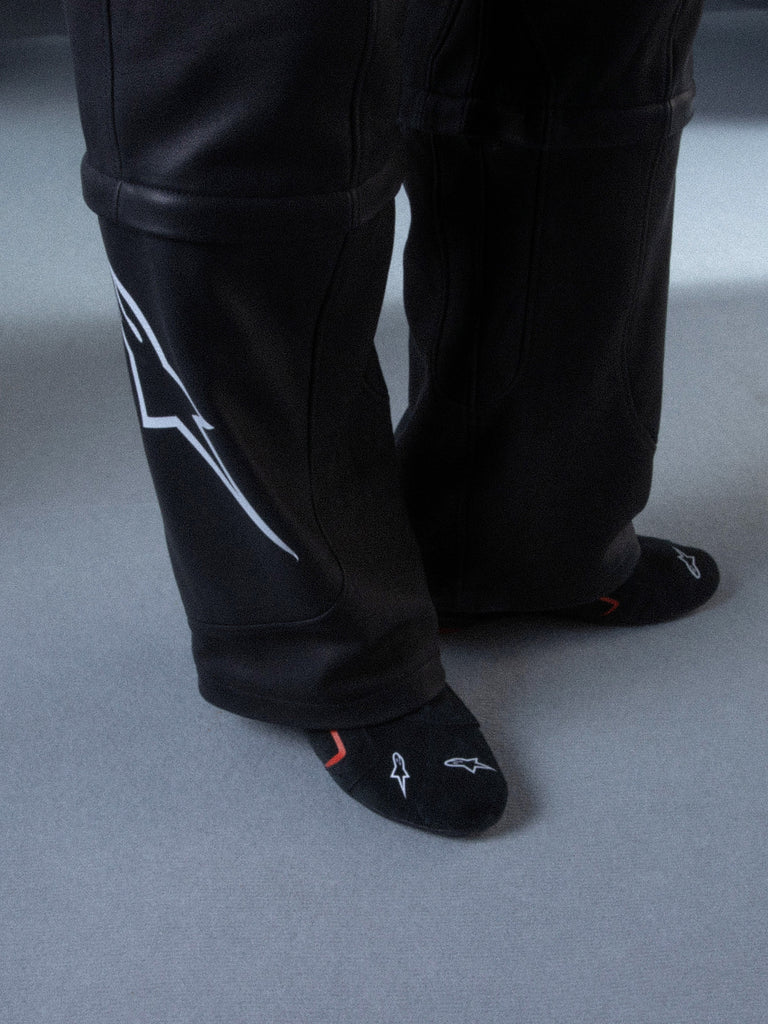 Alpinestars RSRV Starline Shoes, Black premium sneakers with white logo detailing, shown with black socks featuring red and white accents, lifestyle shot on light gray floor