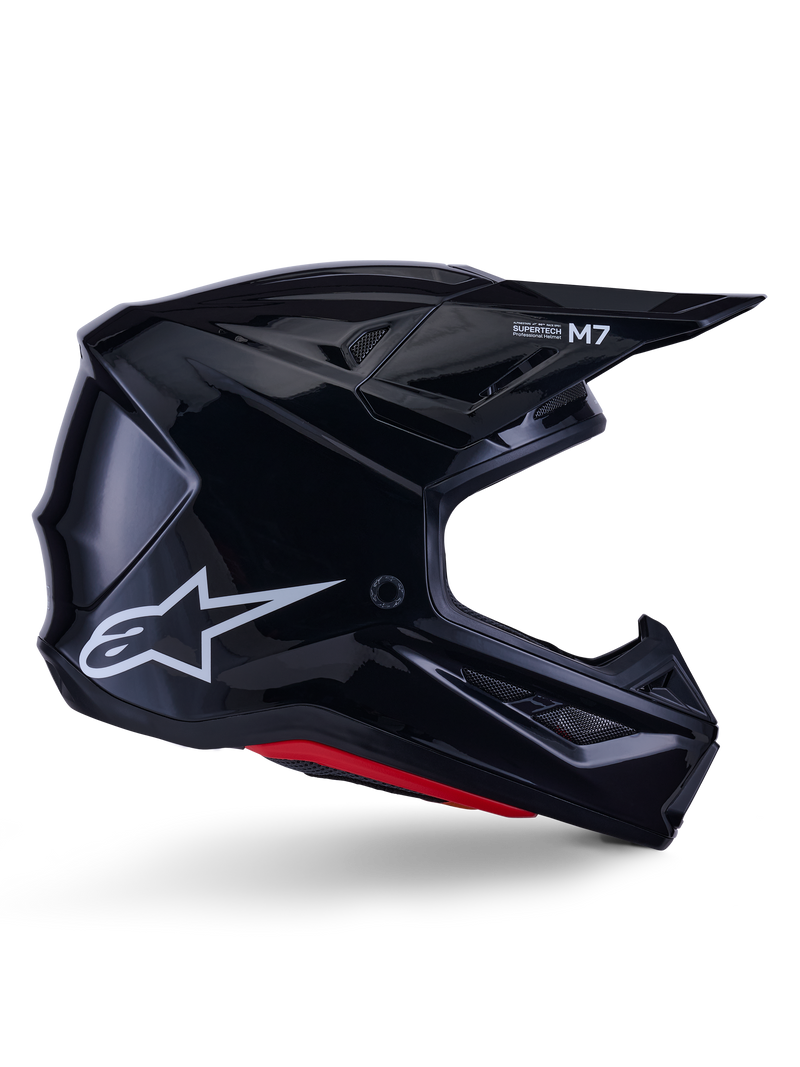 Kask Alpinestars S-M7 Solid, kask motocrossowy, czarny z połyskiem, białe logo gwiazdy i czerwone akcenty, profil boczny prezentujący aerodynamiczny design z kanciastymi otworami wentylacyjnymi i brandingiem Supertech M7 na daszku.