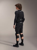 Model w ochraniaczach kolan i goleni Alpinestars A-Impact Plasma Pro, widok od tyłu, kolor czarno-biały, prezentujący potrójne zapięcie na paski i wentylowane panele z siatki, ubrany w pełny zestaw czarnej odzieży ochronnej MTB z koszulką z długim rękawem i spodenkami ochronnymi