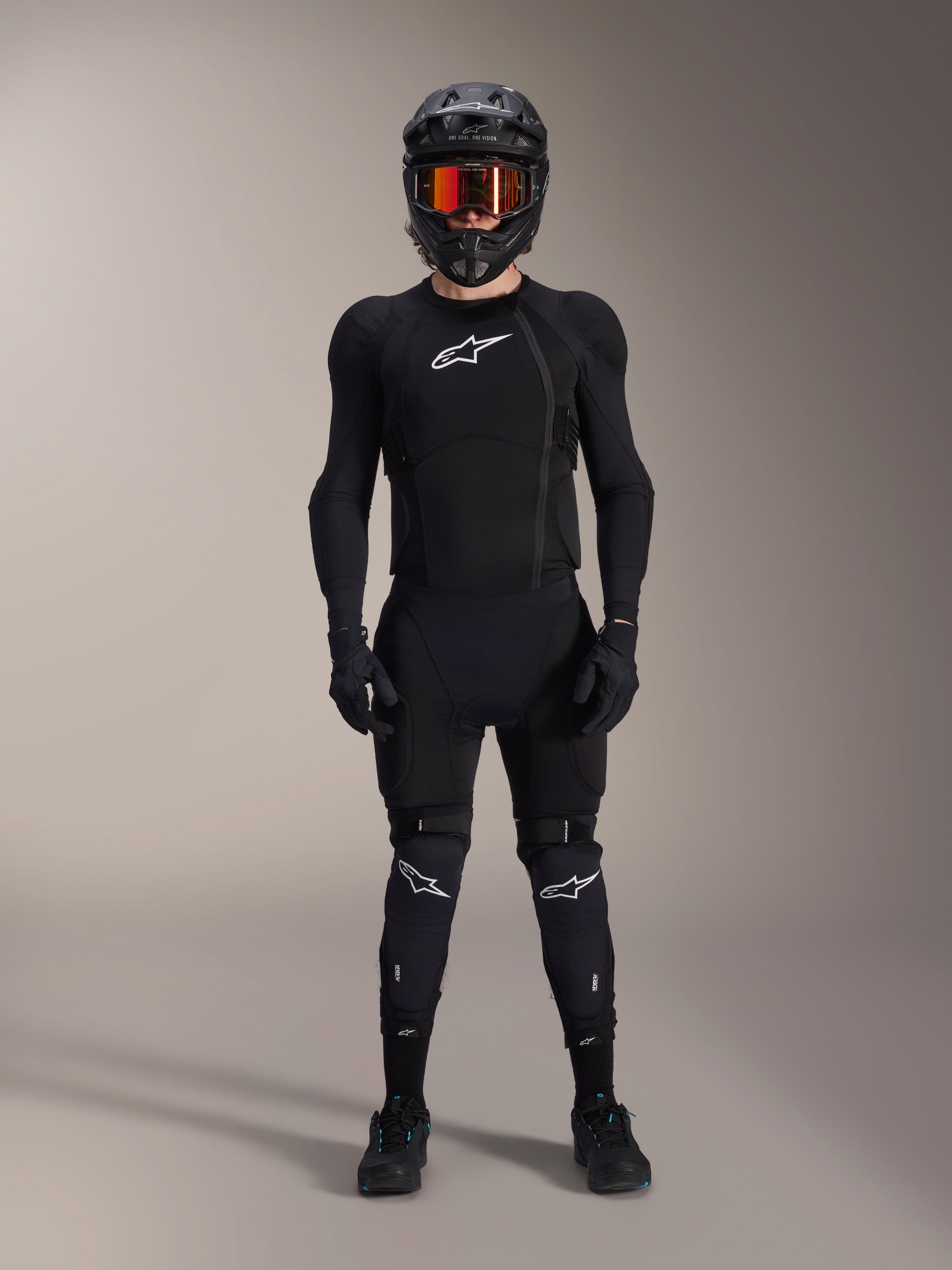 Rowerzysta MTB w pełnym zestawie ochronnym, kurtka ochronna Alpinestars A-Impact Plasma w kolorze czarno-białym, białe logo na klatce piersiowej i siatkowy materiał, czarny kask, gogle z czerwonymi szybkami i ochraniacze kolan, profesjonalny komplet ochraniaczy rowerowych