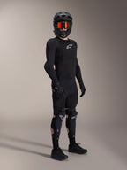 Alpinestars A-Impact Plasma Pro Knee Shin Protectors, czarno-białe, noszone przez rowerzystę w kompletnym stroju ochronnym MTB, zawierającym czarną zbroję z długim rękawem, czarne rękawice oraz kask typu full-face z pomarańczowymi goglami.
