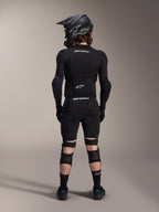Alpinestars A-Impact Plasma Pro Knee Shin Protectors, czarne, widok od tyłu na rowerzystę w pełnym zestawie ochronnym MTB, w tym kasku typu full-face i zbroi, wyposażone w system potrójnego zapięcia na paski oraz wentylowane panele z siatki dla stabilnego dopasowania i przepływu powietrza