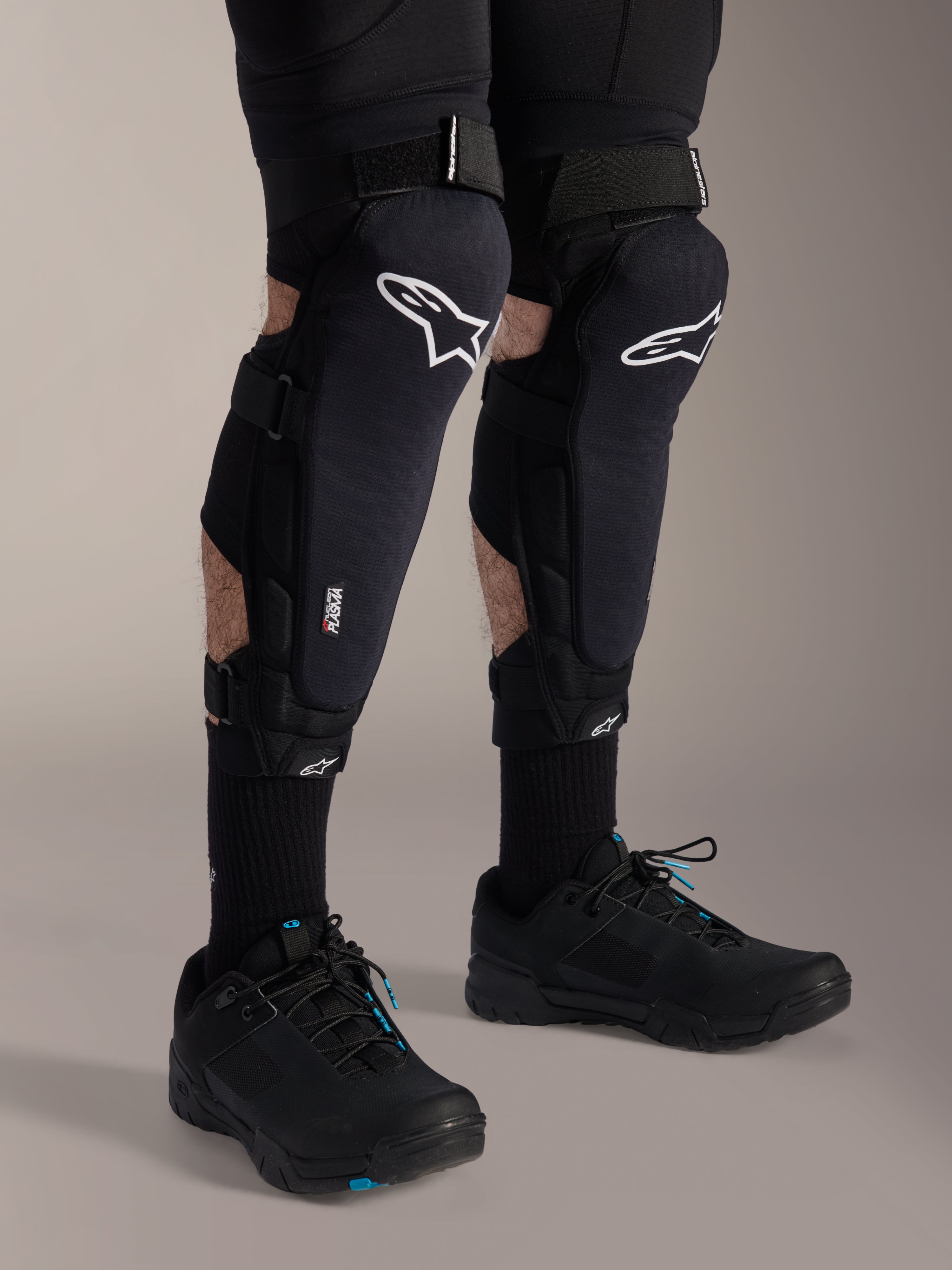 Alpinestars A-Impact Plasma Pro Knee Shin Protectors, czarno-białe, z białym logo Alpinestars i potrójnym systemem zamykania na paski, noszone z czarnymi skarpetami i czarnymi butami sportowymi, wytrzymała ochrona do jazdy terenowej
