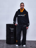 Alpinestars RSRV Arena Suit Jacket, kurtka motocyklowa McLaren Racing, czarna z pomarańczową podszewką, biały napis McLaren Racing, premium streetwear, osoba nosząca kurtkę przy czarnej metalowej beczce