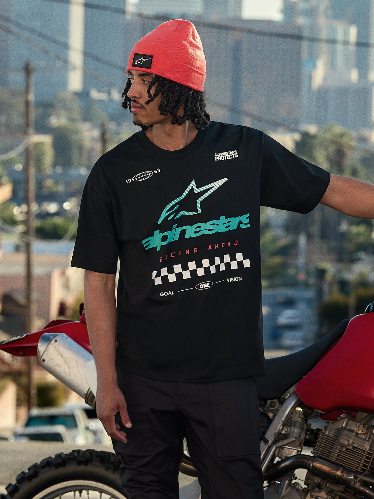 Alpinestars Aplomb Oversized CSF Tee, Short Sleeve T-Shirt, Black, featuring a large teal and white color-gradient Astars logo and checkered flag graphic, worn with a coral beanie, standing next to a red motorcycle, casual lifestyle streetwear
