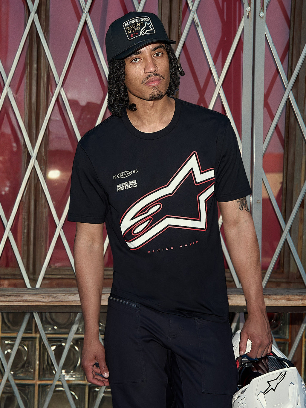 Koszulka Alpinestars Authority CSF, czarny T-shirt z krótkim rękawem, duże biało-czerwone konturowe logo Astars z grafiką „Alpinestars Protects”, ujęcie lifestyle z mężczyzną w czarnej czapce typu trucker trzymającym biały kask, casualowy streetwear motorsportowy