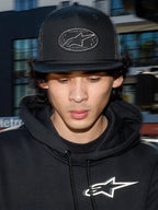 Alpinestars Contender Trucker Hat, czarna czapka lifestylowa z tyłem z siateczki poliestrowej, strukturalna korona z haftowaną naszywką z logo Astars, noszona przez mężczyznę w czarnej bluzie z kapturem Alpinestars, codzienna odzież streetwearowa inspirowana sportami motorowymi.
