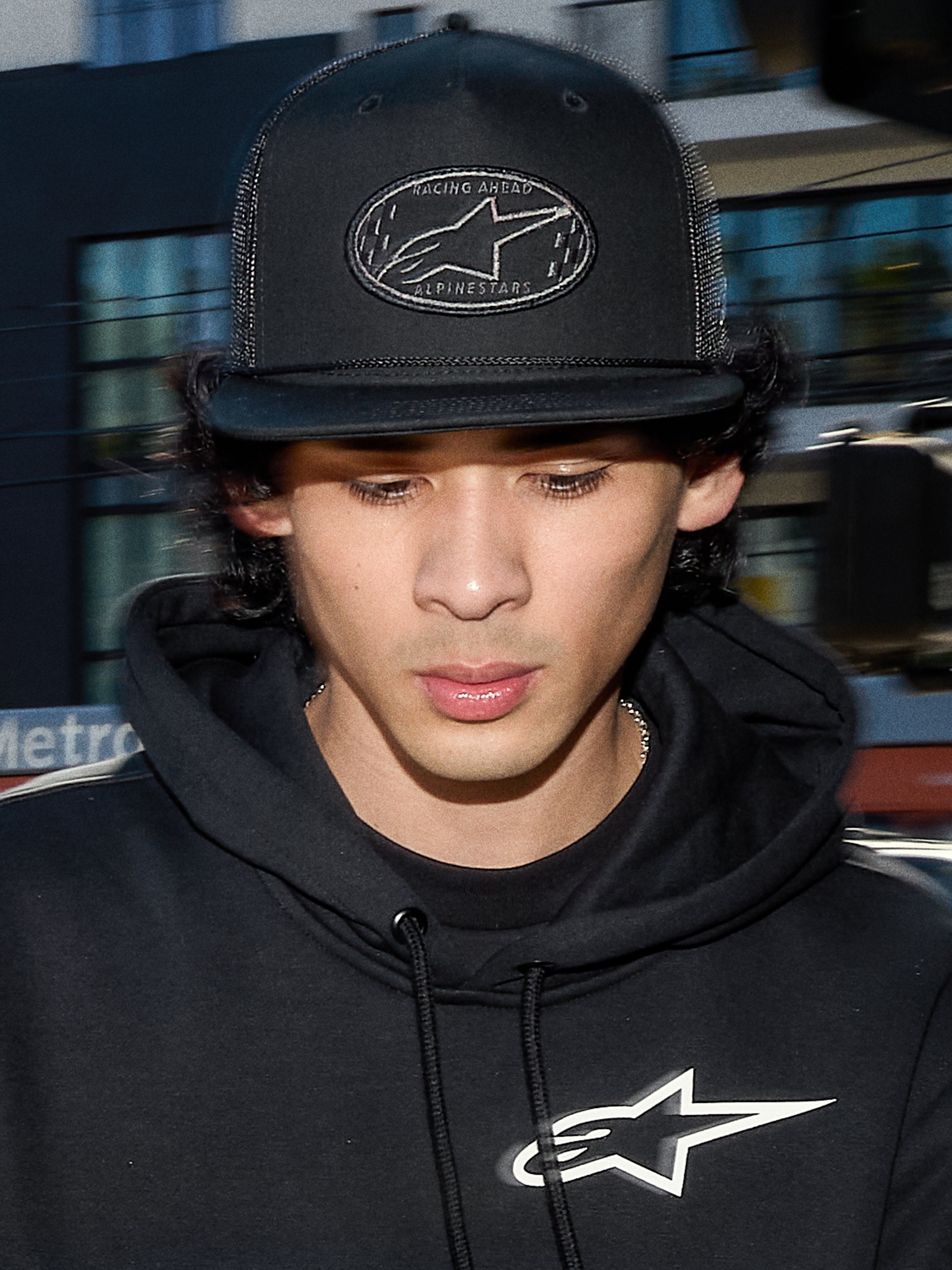 Alpinestars Contender Trucker Hat, czarna czapka lifestylowa z tyłem z siateczki poliestrowej, strukturalna korona z haftowaną naszywką z logo Astars, noszona przez mężczyznę w czarnej bluzie z kapturem Alpinestars, codzienna odzież streetwearowa inspirowana sportami motorowymi.