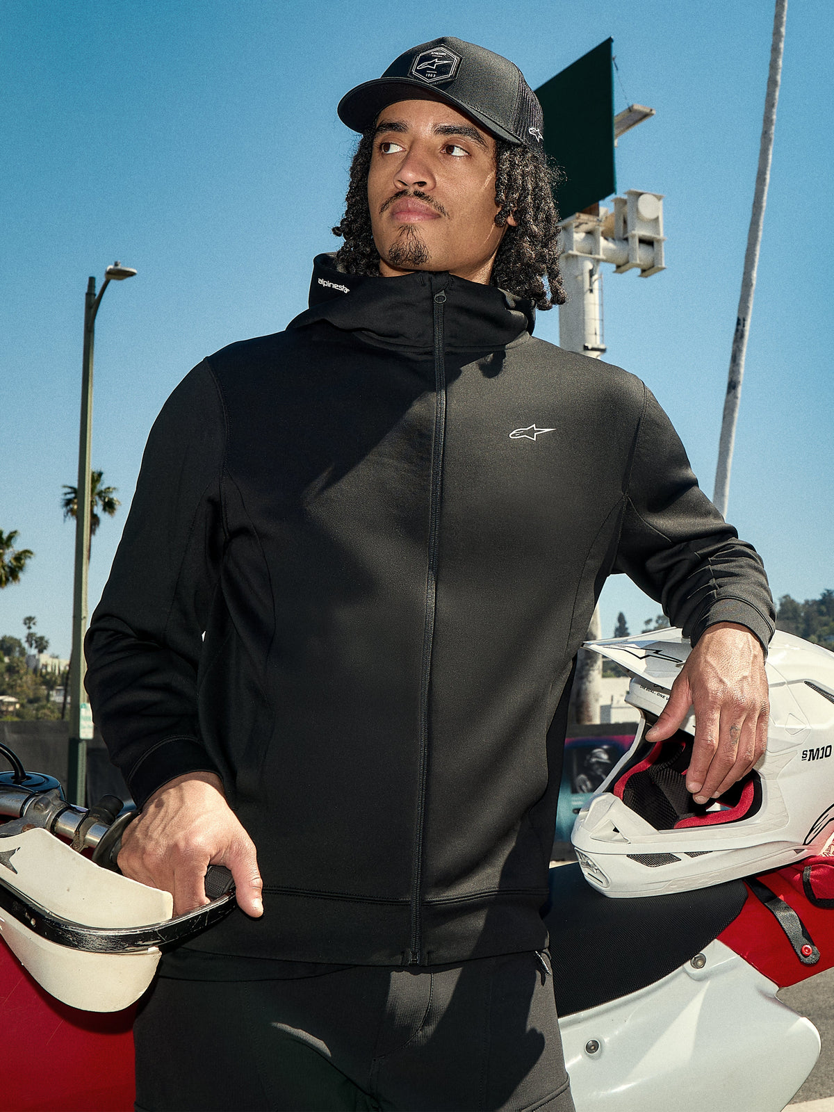 Alpinestars Cypher Hoodie, czarna rozpinana bluza polarowa typu lifestyle, wysoki kaptur z logo marki, małe białe logo Astars na piersi, motocyklista w czarnej czapce trzymający biały kask motocrossowy, sportowy styl streetwear