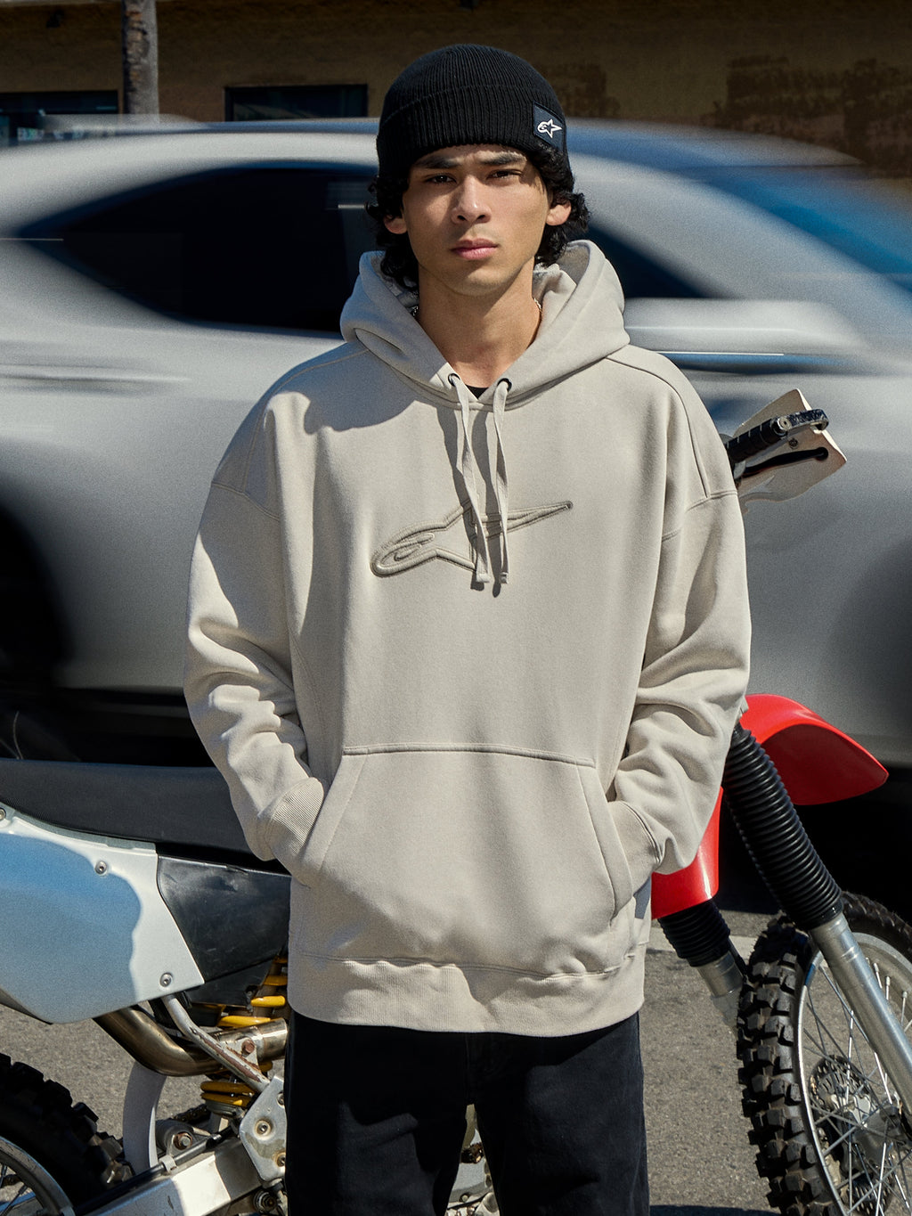 Bluza z kapturem Alpinestars Emergent Oversized w kolorze Off White, oversizowa bluza polarowa z haftowanym logo Astars 3D, kieszenią kangurką i kapturem ze sznurkiem, mężczyzna w czarnej czapce beanie obok motocykla, casualowy styl motorsport streetwear