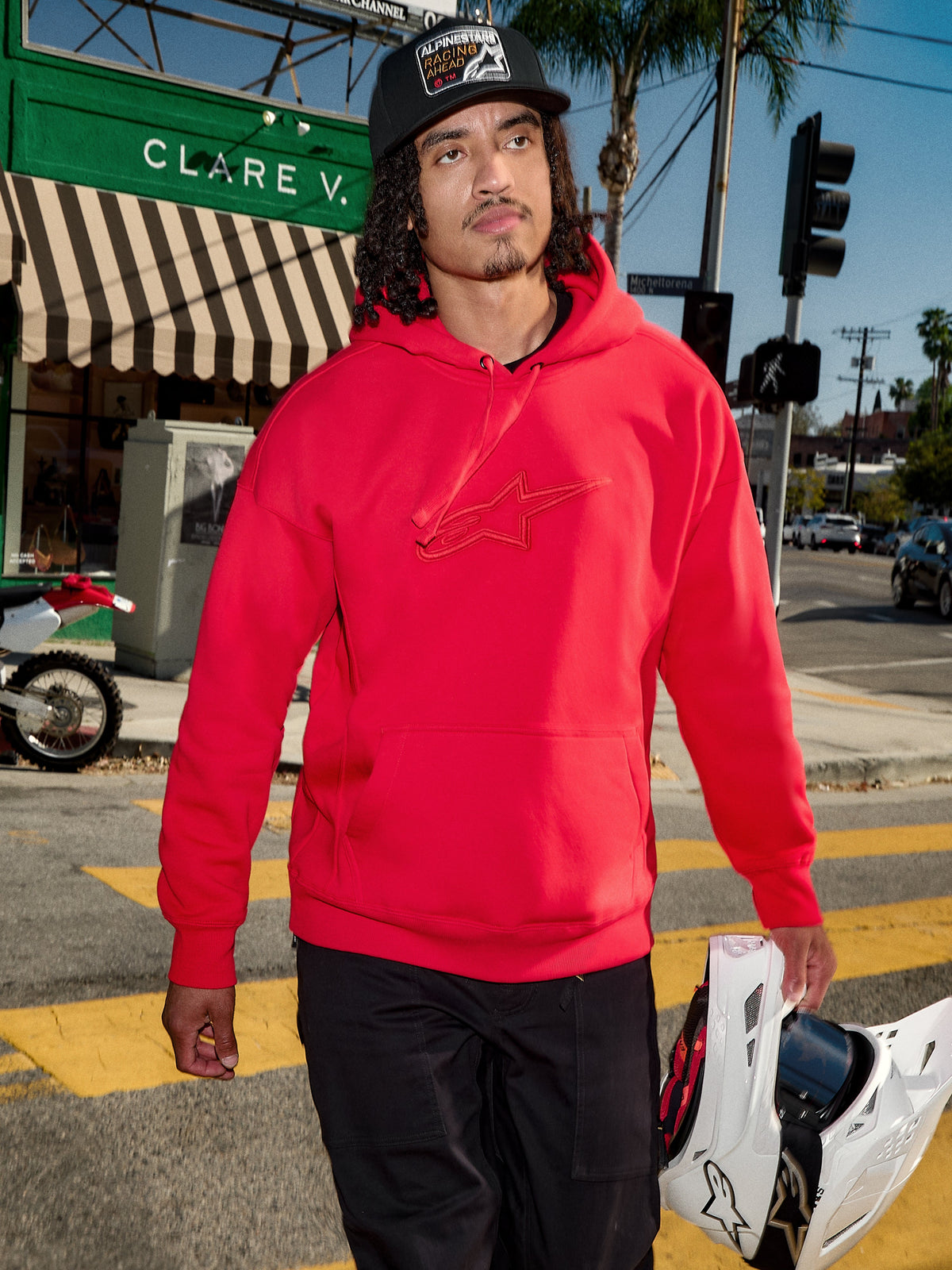 Alpinestars Emergent Oversized Hoodie, czerwona, haftowane logo Astars 3D na piersi, noszona z czarnymi spodniami i czapką z daszkiem, trzymany biały kask motocrossowy, casualowy miejski styl streetwearowy