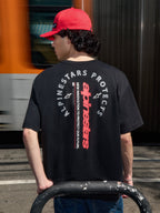 Koszulka Alpinestars Overhang Oversized CSF, czarny T-shirt z krótkim rękawem, widok z tyłu z pionowym czerwonym logotypem i okrągłą grafiką Alpinestars Protects, noszona z czerwoną czapką, casualowy styl motorsport streetwear
