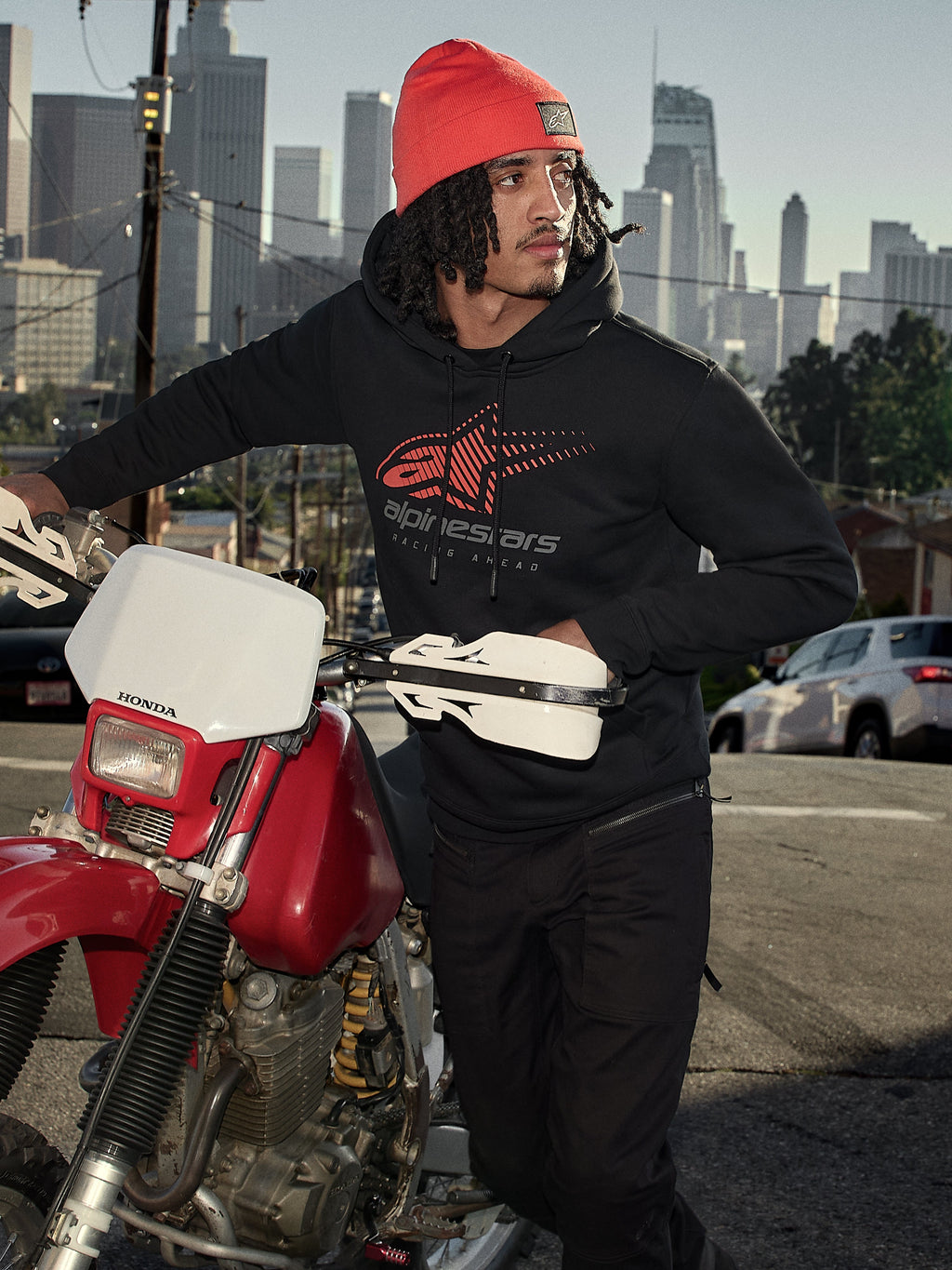 Alpinestars Pulse Hoodie, czarna polarowa bluza z kapturem z czerwono-szarym logo Astars Combo, mężczyzna w pomarańczowej czapce stojący obok czerwonego motocykla Honda w miejskiej scenerii, codzienna odzież motorsport streetwear