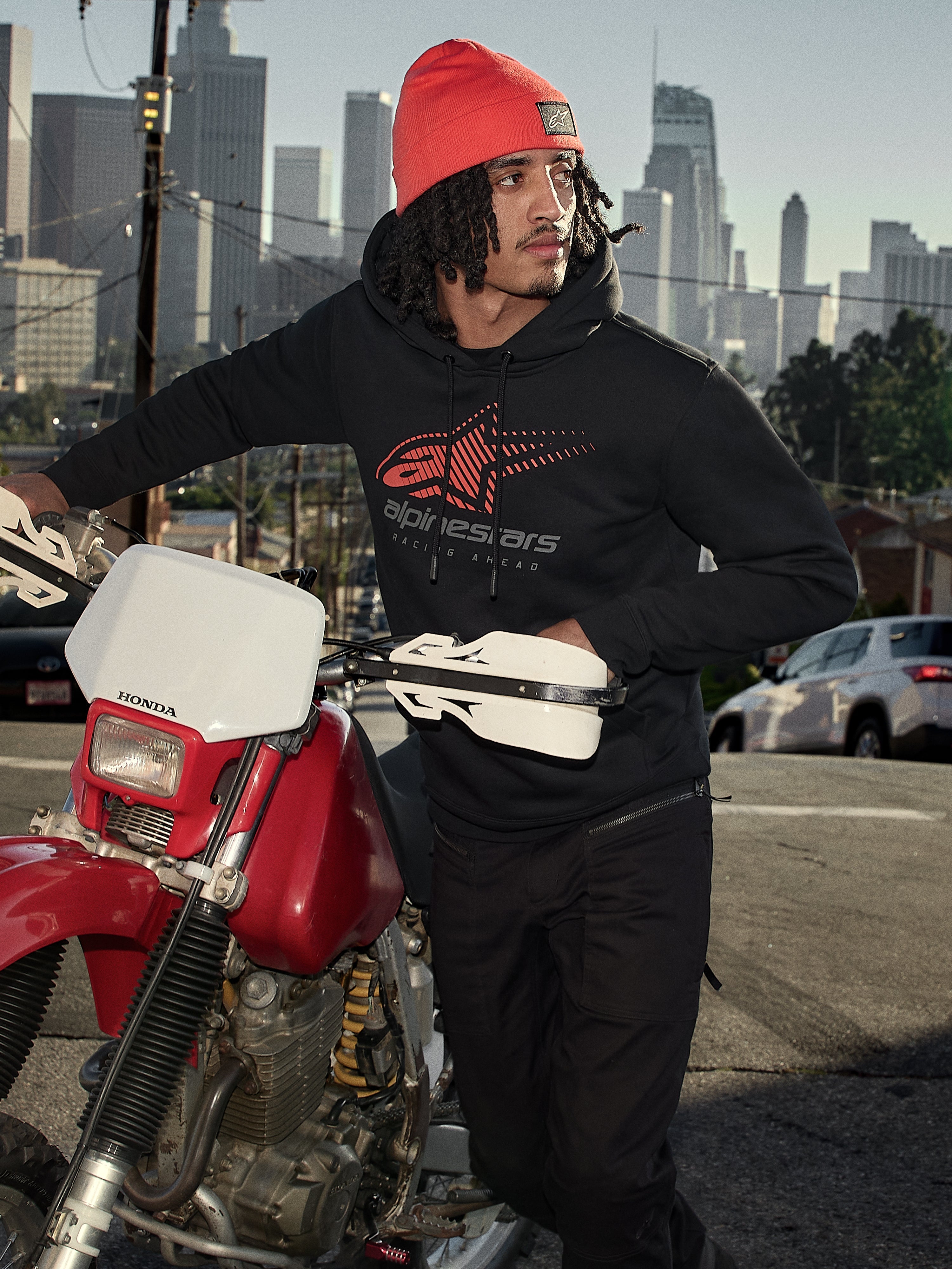 Alpinestars Pulse Hoodie, czarna polarowa bluza z kapturem z czerwono-szarym logo Astars Combo, mężczyzna w pomarańczowej czapce stojący obok czerwonego motocykla Honda w miejskiej scenerii, codzienna odzież motorsport streetwear