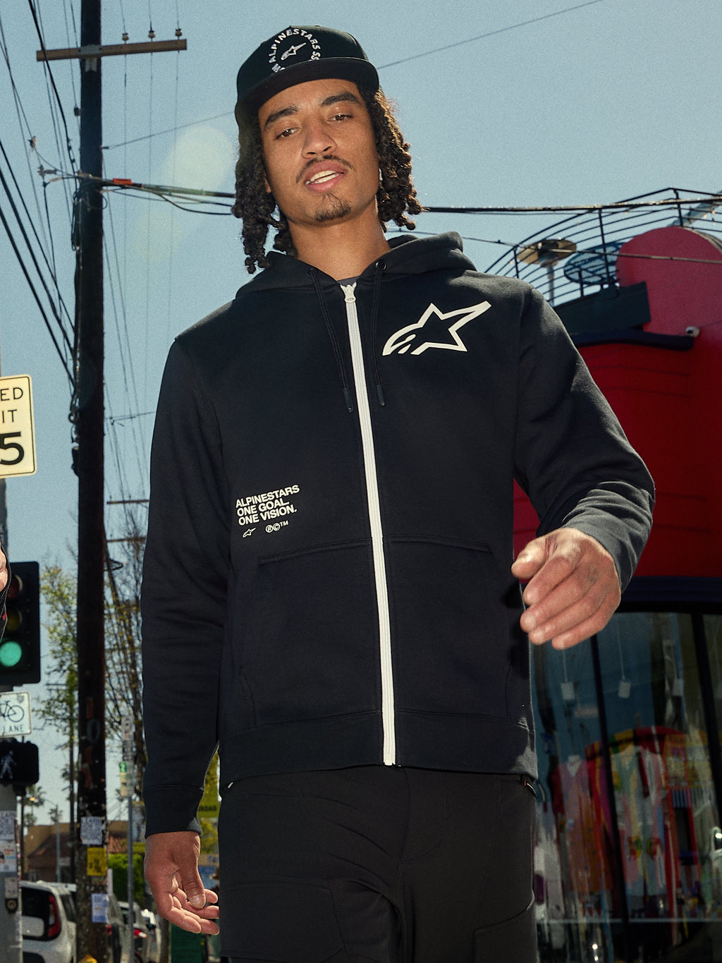 Alpinestars Transverse Hoodie, czarna bluza z polaru zapinana na zamek, biały zamek i białe logo Astars na piersi, nadruk „One Goal One Vision”, codzienny styl streetwear, mężczyzna w miejskim otoczeniu