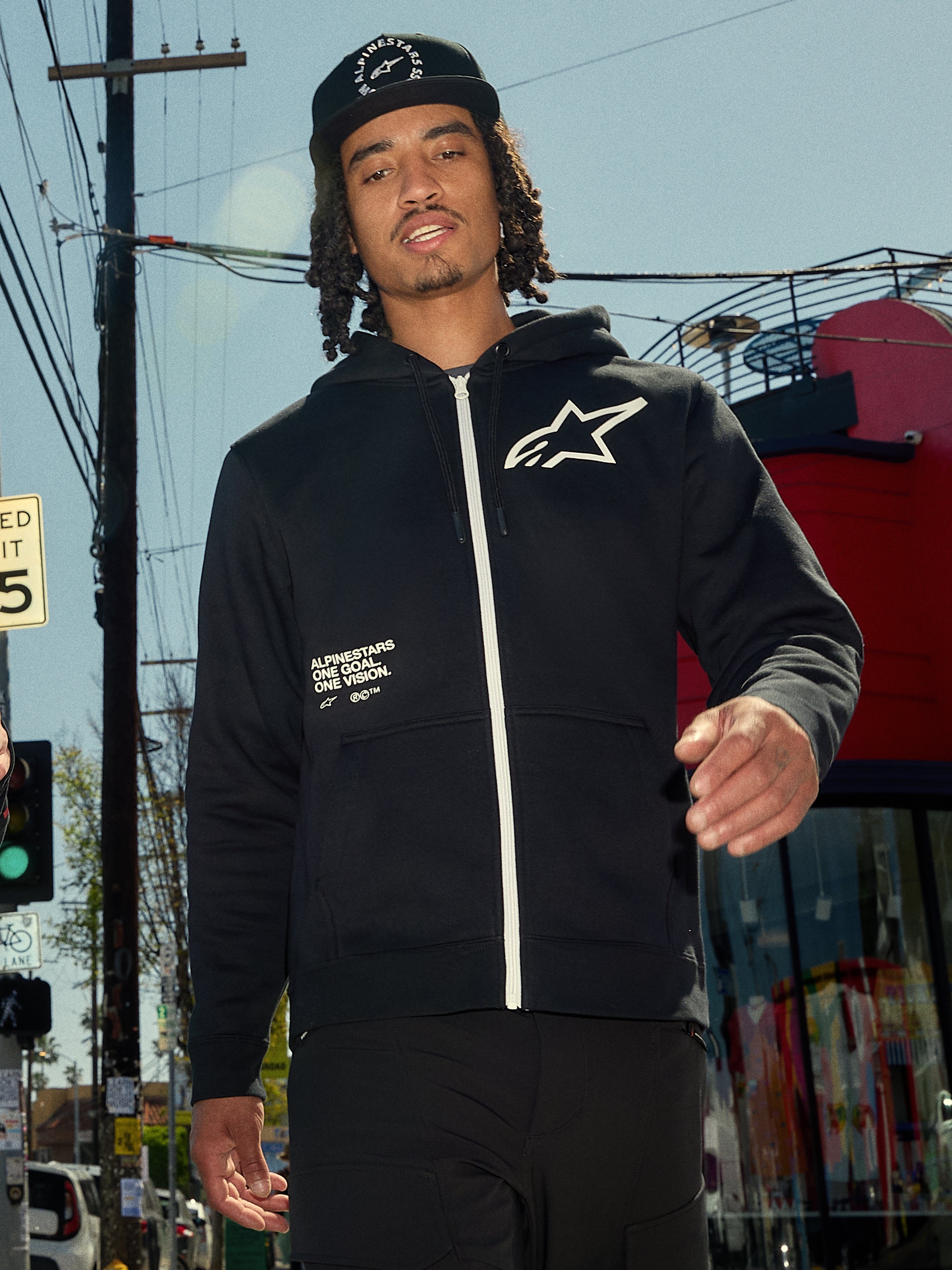 Alpinestars Transverse Hoodie, czarna bluza z polaru zapinana na zamek, biały zamek i białe logo Astars na piersi, nadruk „One Goal One Vision”, codzienny styl streetwear, mężczyzna w miejskim otoczeniu