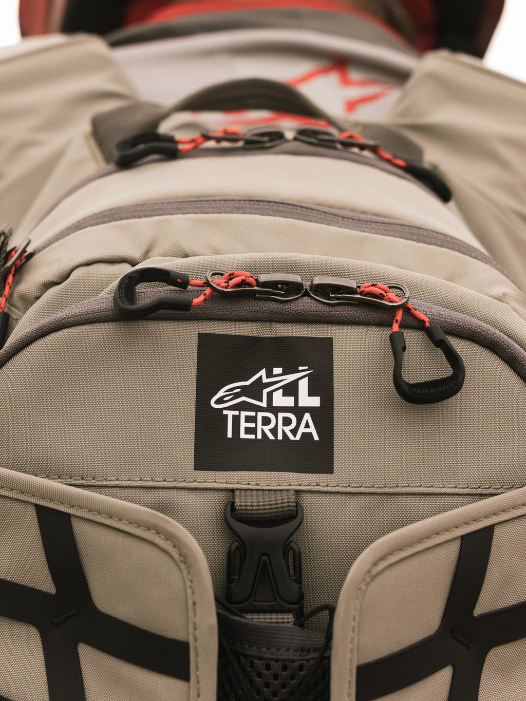 Plecak Alpinestars AT-15, Aluminum Walnut, zbliżenie na naszywkę z logo All Terra na beżowej tkaninie technicznej, pomarańczowe sznurkowe uchwyty zamków, wytrzymałe detale konstrukcyjne