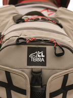 Plecak Alpinestars AT-15, Aluminum Walnut, zbliżenie na naszywkę z logo All Terra na beżowej tkaninie technicznej, pomarańczowe sznurkowe uchwyty zamków, wytrzymałe detale konstrukcyjne