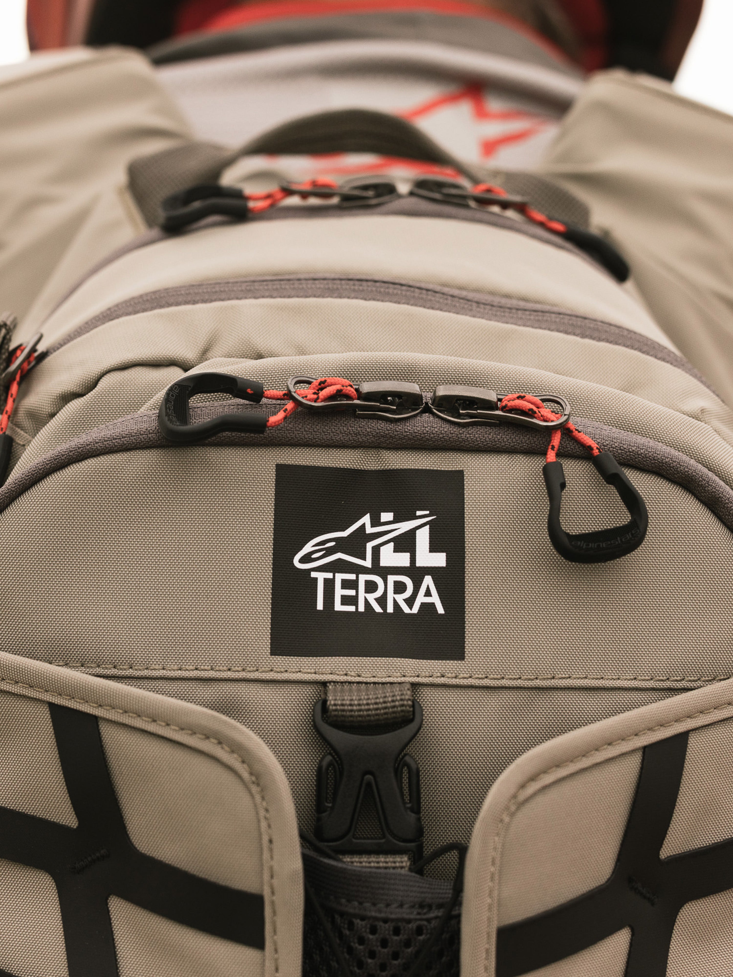 Plecak Alpinestars AT-15, Aluminum Walnut, zbliżenie na naszywkę z logo All Terra na beżowej tkaninie technicznej, pomarańczowe sznurkowe uchwyty zamków, wytrzymałe detale konstrukcyjne