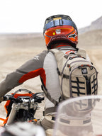 Plecak Alpinestars AT-15 w kolorze Aluminum Walnut, noszony przez motocyklistę adventure w pustynnym krajobrazie, wytrzymała beżowa konstrukcja tekstylna z czarnymi taśmami, pomarańczowymi akcentami przy zamkach i brandingiem „All Terra”, zestawiony z czerwono-pomarańczowym kaskiem i szarą odzieżą motocyklową do turystyki off-road