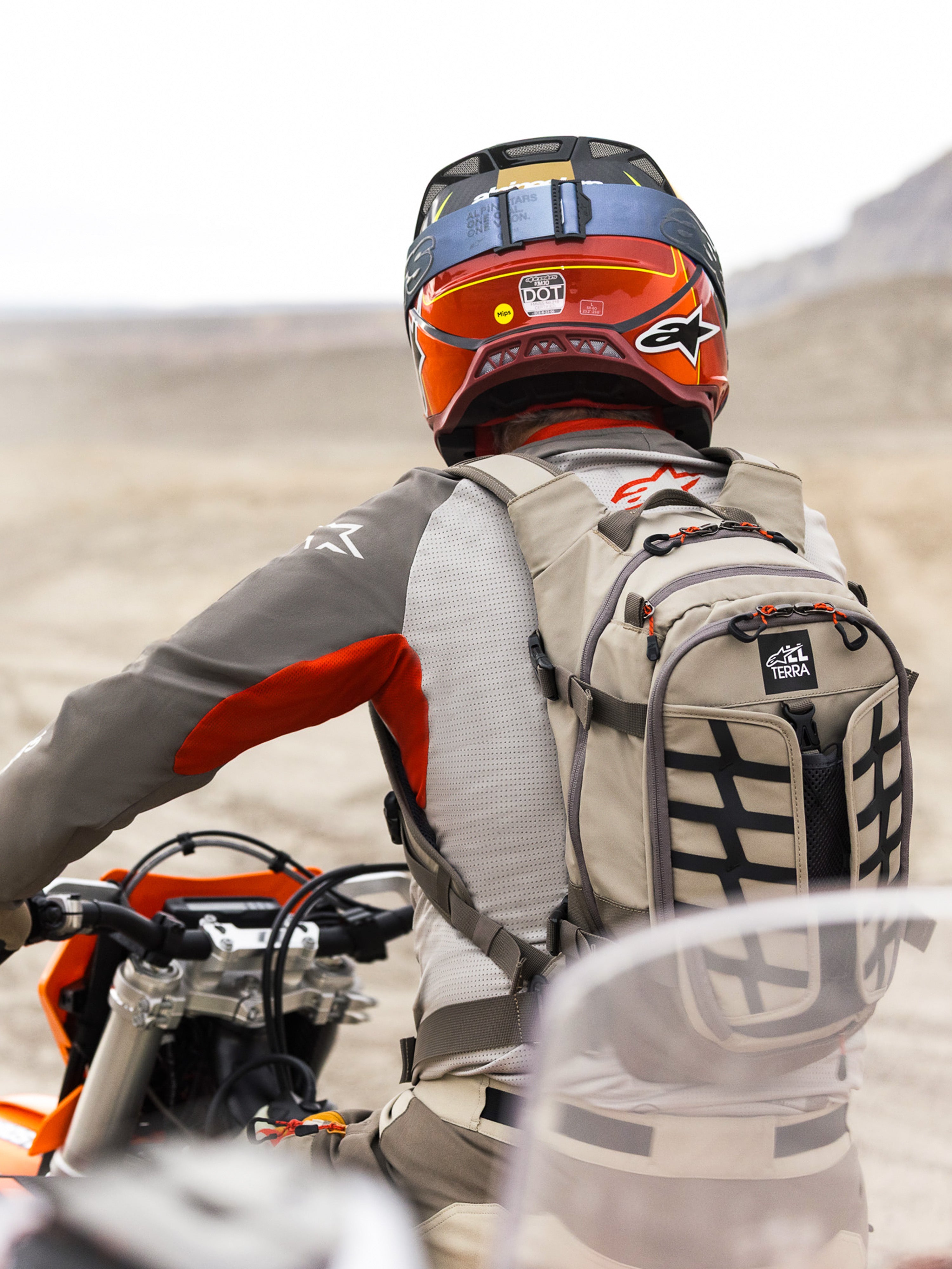 Plecak Alpinestars AT-15 w kolorze Aluminum Walnut, noszony przez motocyklistę adventure w pustynnym krajobrazie, wytrzymała beżowa konstrukcja tekstylna z czarnymi taśmami, pomarańczowymi akcentami przy zamkach i brandingiem „All Terra”, zestawiony z czerwono-pomarańczowym kaskiem i szarą odzieżą motocyklową do turystyki off-road