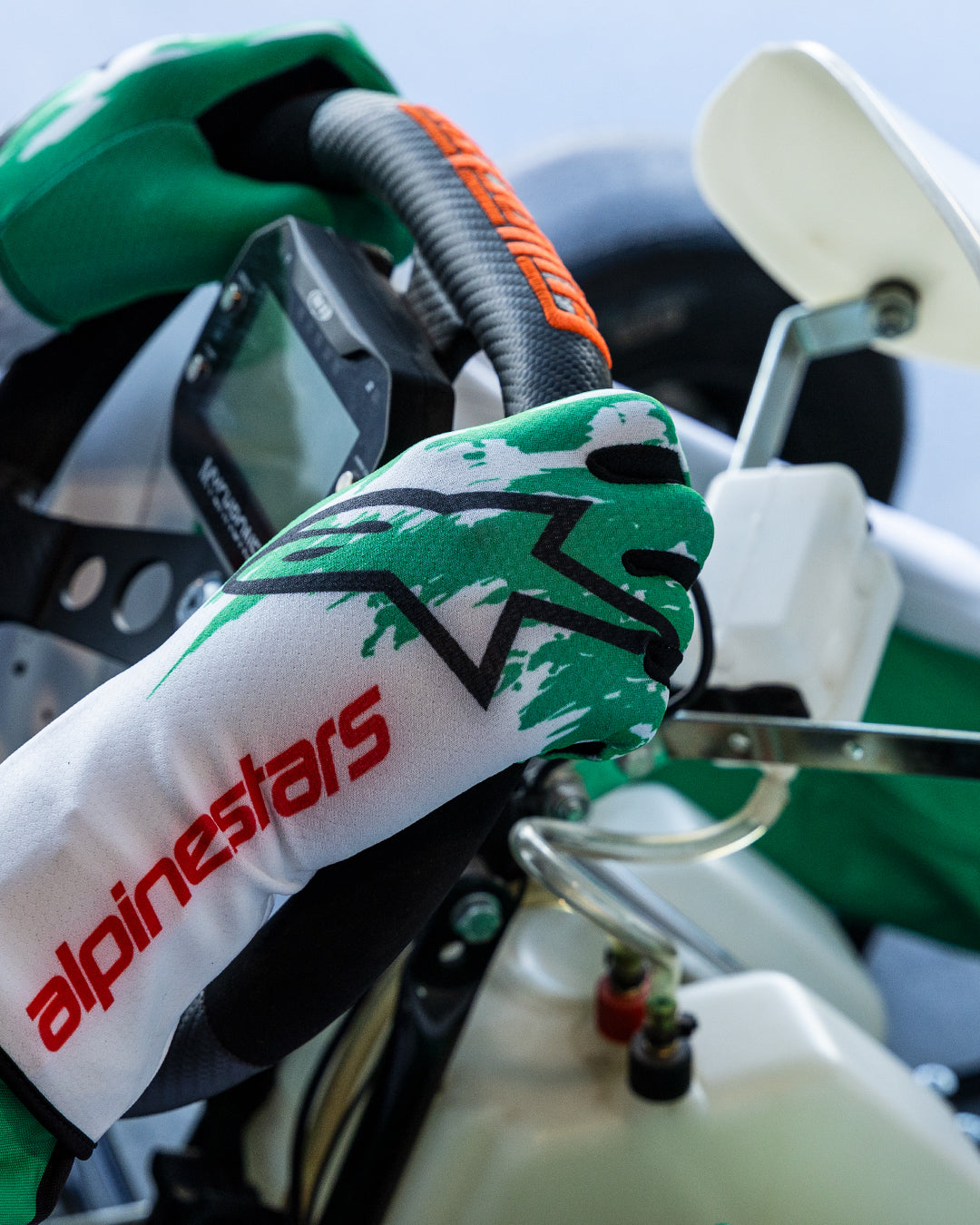 Rękawice Alpinestars Tech-1 K V3 w kolorze biało-zielono-czerwonym, dłonie kierowcy trzymające kierownicę gokarta, zielony sublimowany wzór splash, czarne logo z gwiazdą i czerwony napis Alpinestars na mankiecie, profesjonalny sprzęt kartingowy