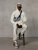 Bluza z kapturem Alpinestars Essentials Ageless, Off-White, noszona przez modela siedzącego na stołku z pasującymi polarowymi joggerami, beżową czapką ASTARS i torbą typu cross-body, casualowy streetwear inspirowany sportami motorowymi