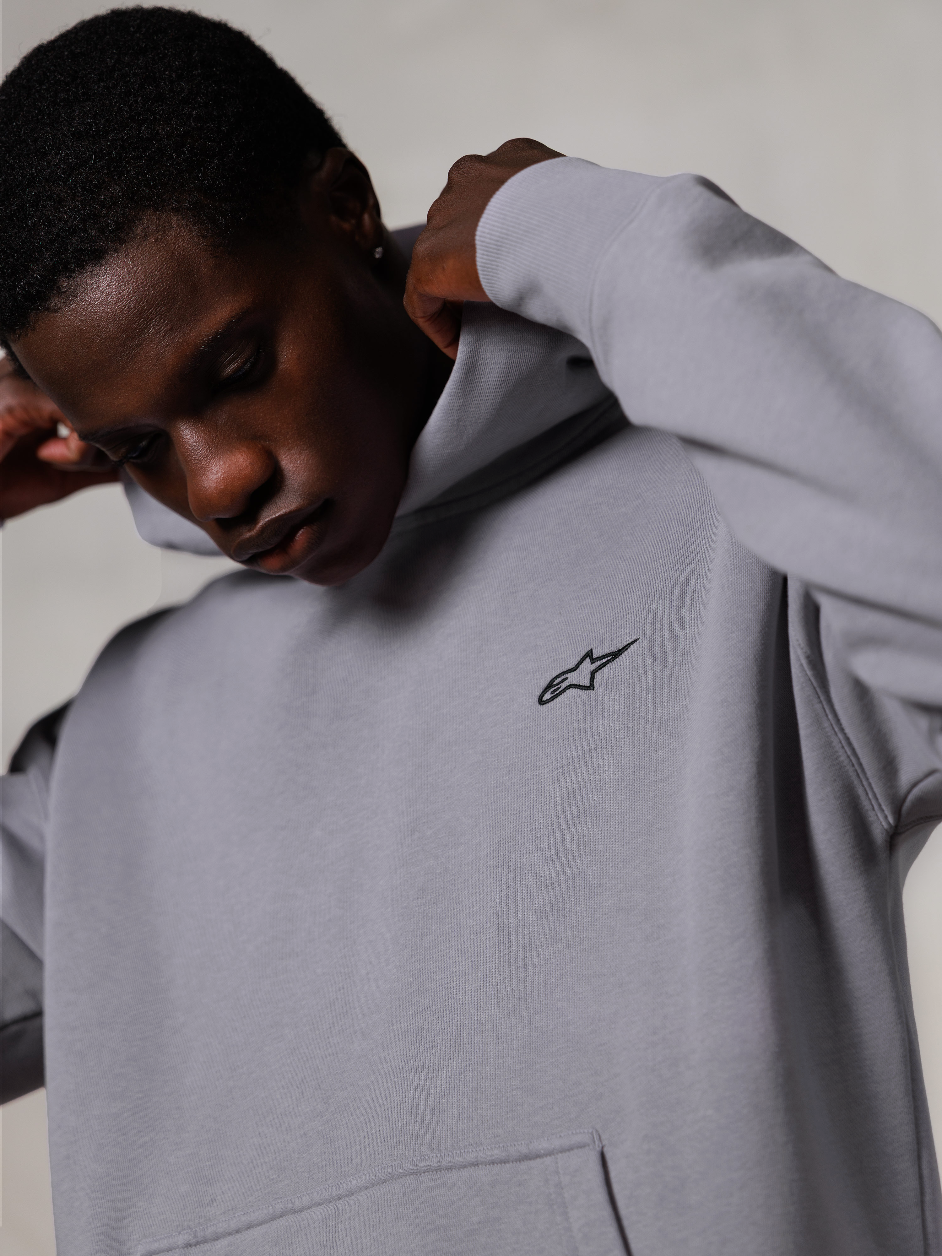 Alpinestars Essentials Ageless Hoodie, srebrna bluza z kapturem, zbliżenie na mężczyznę poprawiającego kaptur szarej bluzy, małe czarne logo Alpinestars na piersi, odzież codzienna premium inspirowana sportami motorowymi