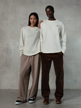 Alpinestars Essentials Ageless LS Tee, koszulka z długim rękawem w kolorze złamanej bieli, prezentowana przez modelkę i modela, małe czarne logo Alpinestars na piersi, luźny krój, stylizacja z beżowymi spodniami dresowymi i brązowymi spodniami sztruksowymi, casualowy streetwear inspirowany sportami motorowymi